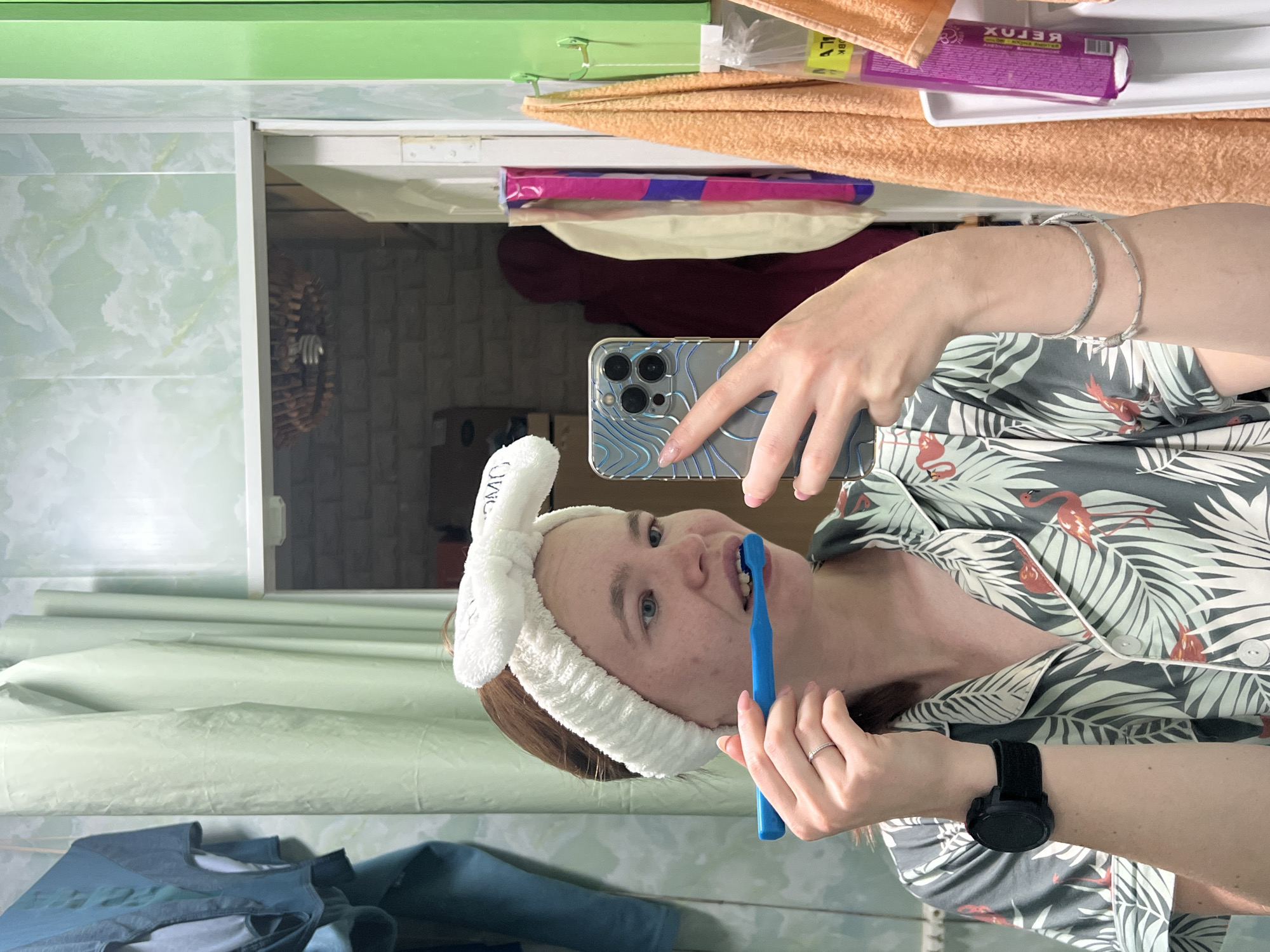 In the city of Chaikovsky, Russia, a young woman with auburn hair, wearing a white plush headband and a pajama top with a tropical print featuring flamingos and palm leaves, is captured in a mirror selfie. She is in a bathroom setting, evident from the tiled walls and the presence of various toiletries and towels. The woman is holding a blue toothbrush in her right hand, applying it to her teeth, suggesting she is brushing them. Her left hand is holding a smartphone with a decorative, iridescent case, angled to capture the reflection. She is wearing a black smartwatch on her left wrist and two thin bracelets on her right arm. The bathroom appears to be brightly lit, with a green shower curtain and a partially open door revealing a brick-patterned wall in the background. Several towels, one orange and one light peach, hang on hooks to the right of the frame. A small object, possibly a cleaning brush head, is visible hanging from a hook above the doorway.
