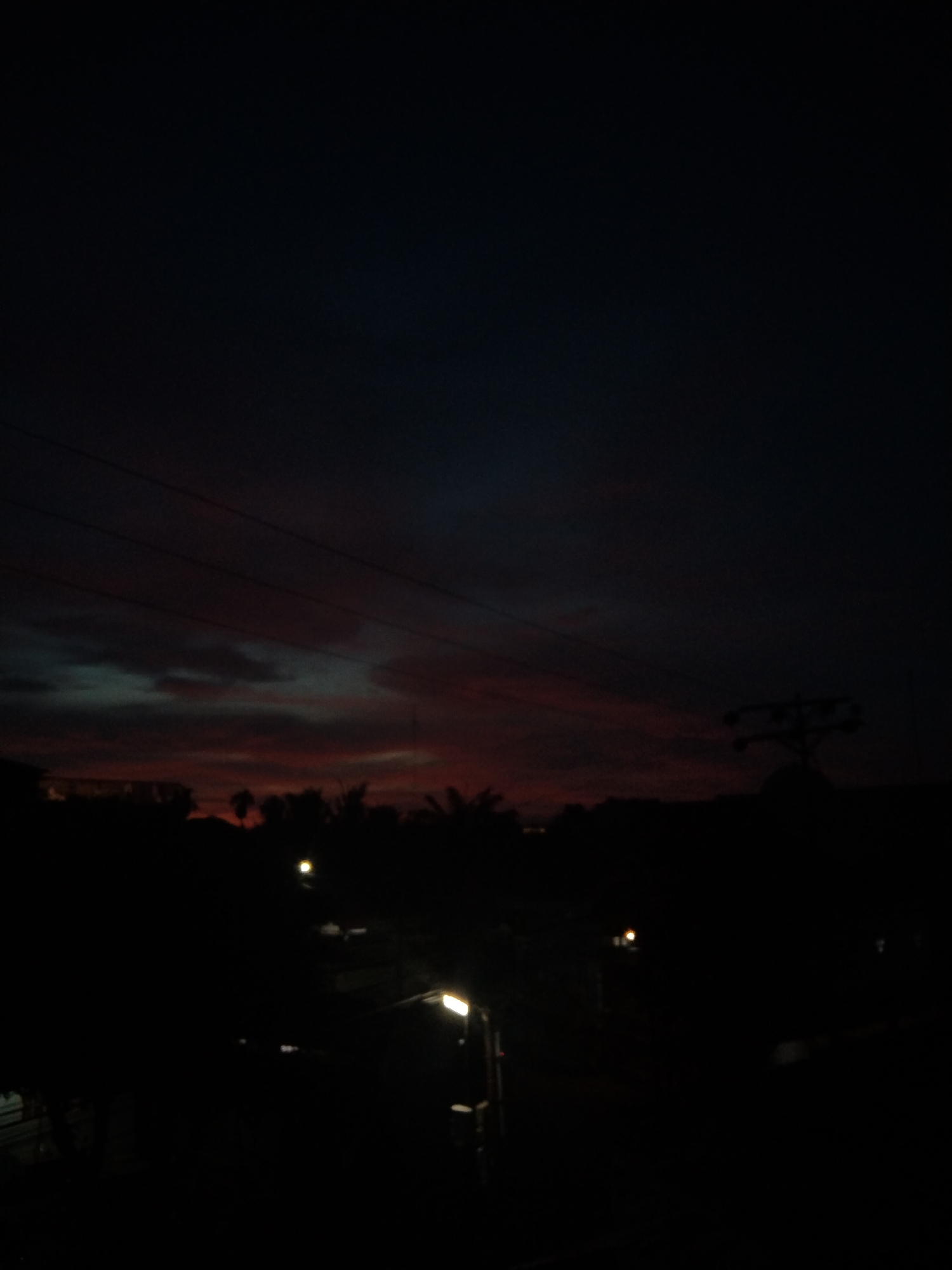 The image depicts a nighttime scene in Bireuenmeunasahdayah, Indonesia. A dark sky with streaks of red and gray clouds is visible above the horizon. Silhouettes of trees and buildings are visible along the horizon line. Several streetlights illuminate the scene from below. Power lines stretch across the upper portion of the image. A utility pole is on the right.
