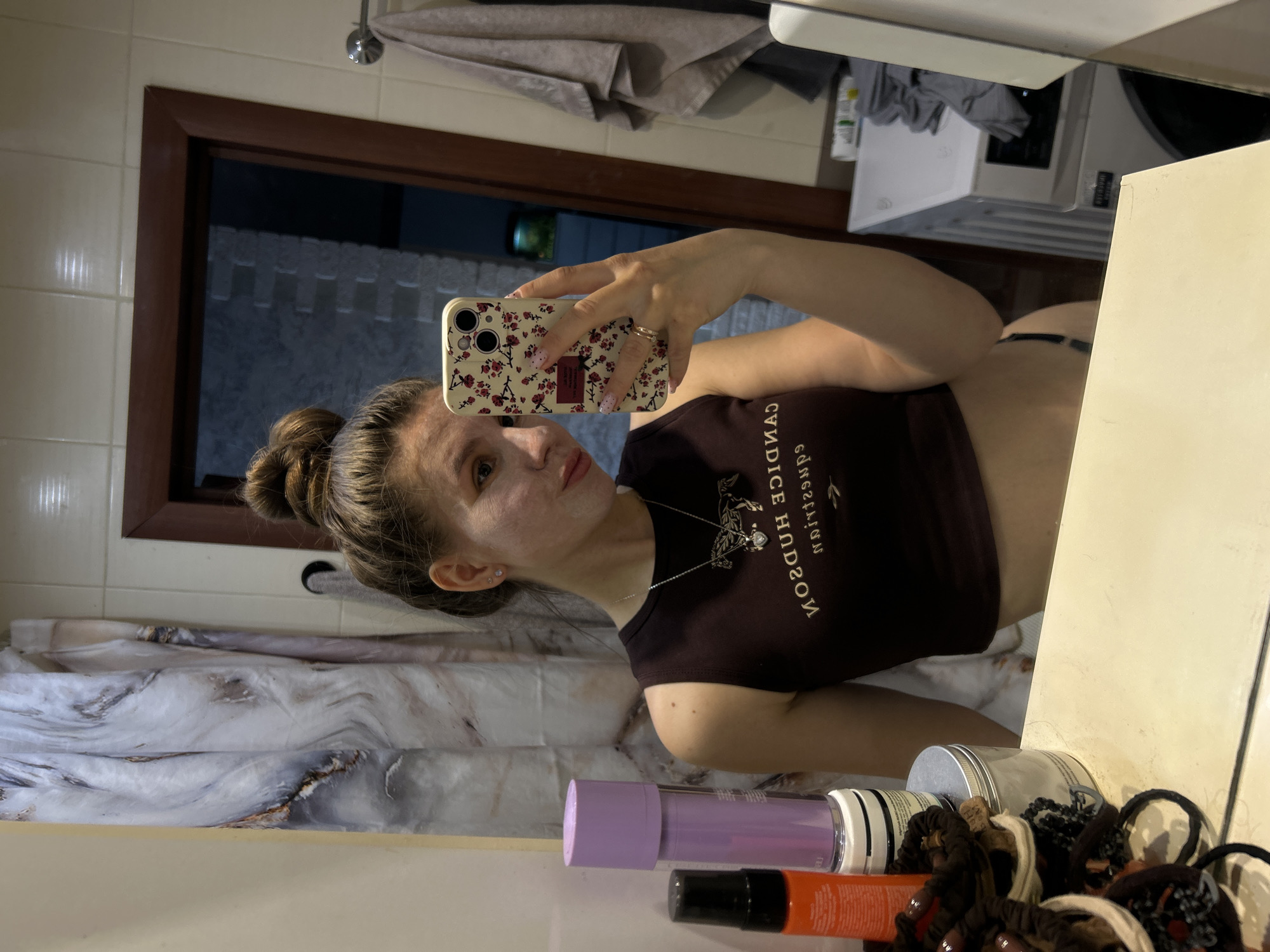 A woman is taking a selfie in a bathroom with a brown-framed mirror, white tiled walls, and a marble-patterned shower curtain. Her hair is styled in a bun. She is wearing a brown crop top and a necklace. Her phone has a patterned case. On the bathroom counter are various hair and beauty products. A washing machine is visible in the background. The location is implied to be Klin, Russia.