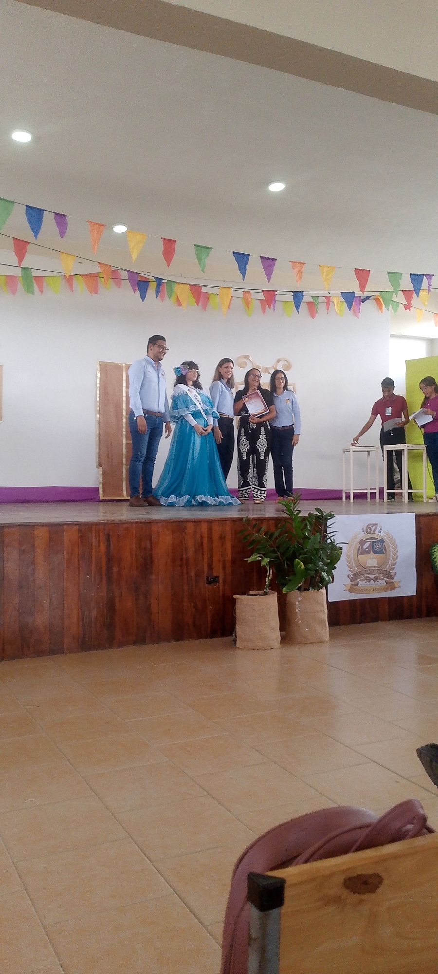 A photograph taken indoors shows a stage on which a group of people stand. The scene takes place in Juan Griego, Venezuela. The stage is decorated with colorful triangular flags hung from the ceiling. A man stands to the left wearing a light blue shirt and jeans. Next to him stands a young woman wearing a blue dress, a sash, and a floral headpiece. Three other women stand next to her, all wearing light blue shirts. The woman in the middle holds a document. To the right of the group, two people are standing near a small white table. A sign with a logo is placed on the front of the stage near potted plants. The floor is tiled, and chairs are visible in the foreground.