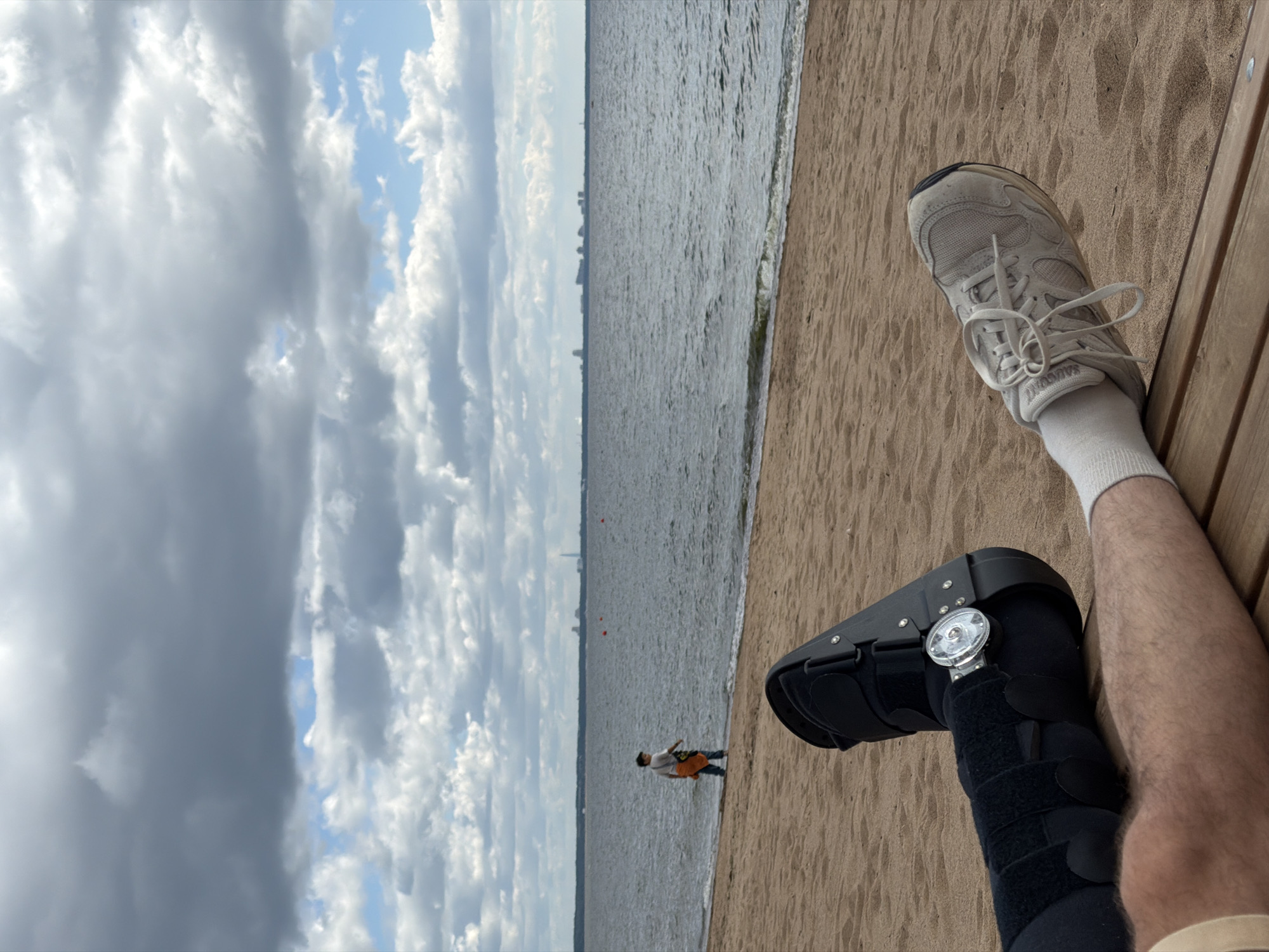 This is a close-up, eye-level, medium shot of a person's legs and feet resting on a wooden bench at a beach. The person is wearing a walking boot on their left leg and a light gray sneaker on their right foot, along with white socks. The left boot is black with Velcro straps and has a silver dial adjustment mechanism on the side. The background shows a sandy beach, a body of water, and a cloudy sky. In the distance, along the shoreline, a person is walking, and a city skyline with tall buildings and industrial structures is visible on the horizon. The overall setting appears to be a public beach in a coastal area.