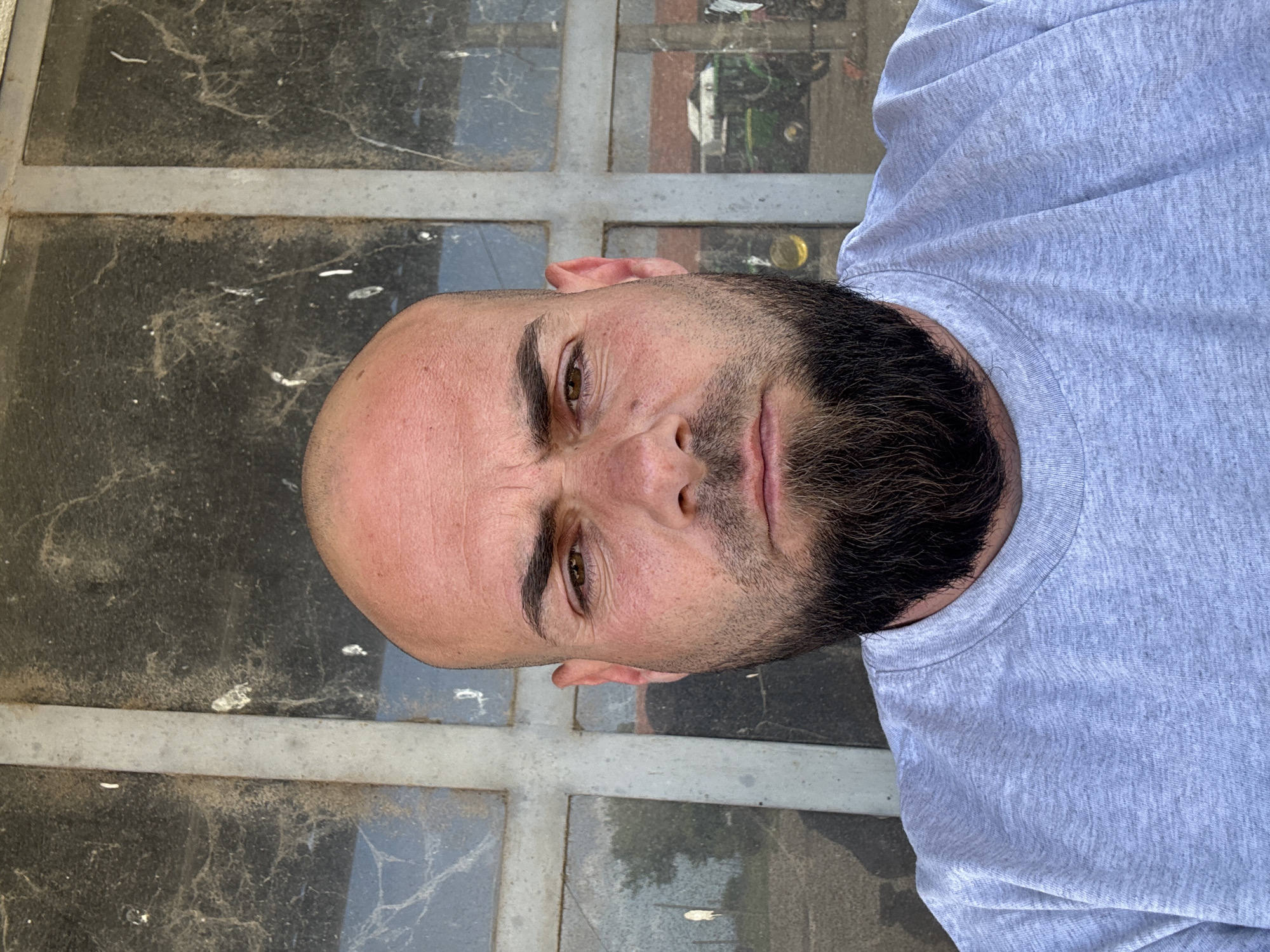 The image shows a bald man with a dark beard wearing a gray t-shirt. He appears to be outdoors, standing in front of a window or surface covered in dust and spiderwebs. It is impossible to provide a location in the city of Sarnevo, Bulgaria with the given information.