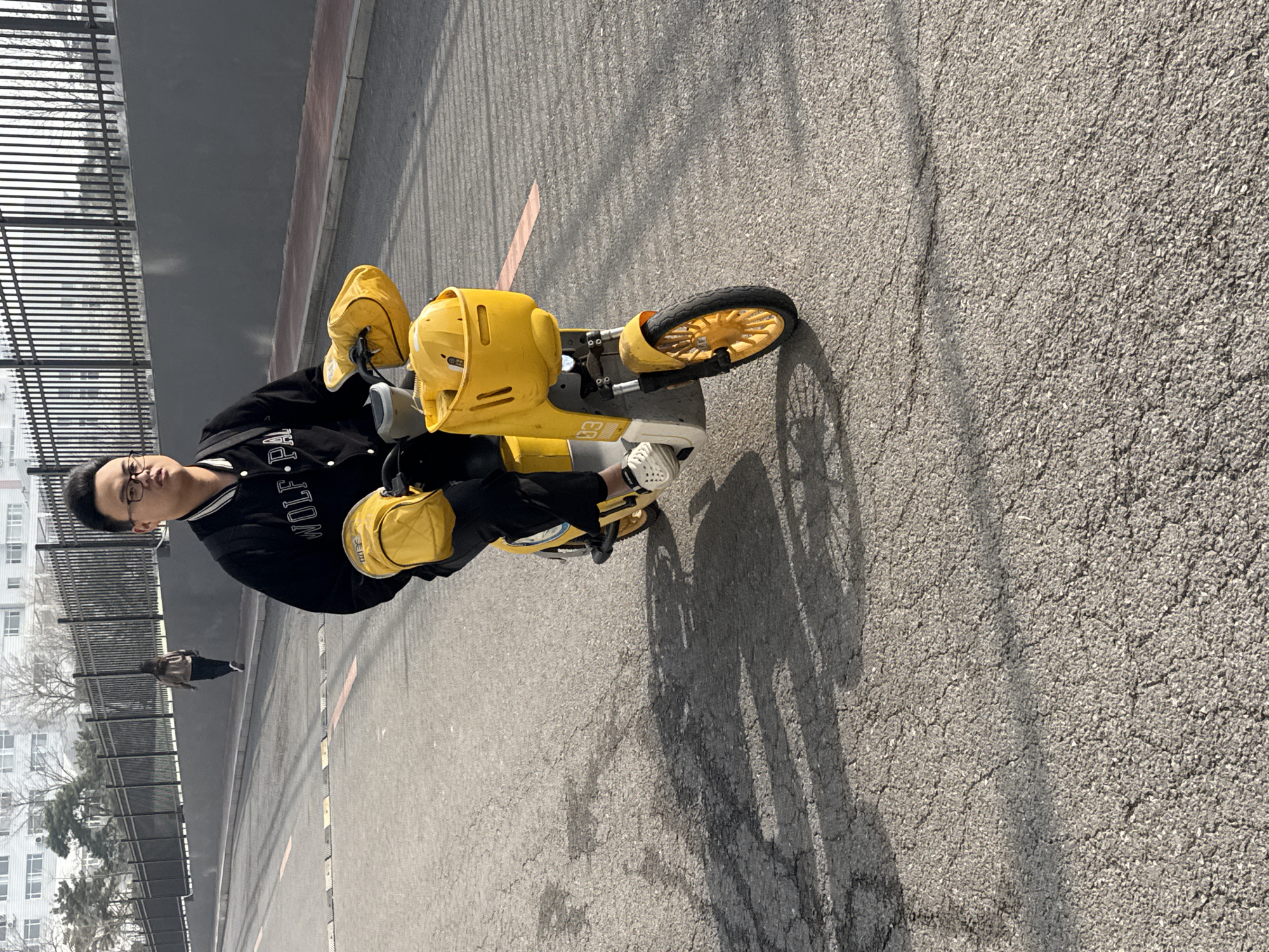 A man with glasses is sitting on a small yellow bicycle in Zhongtun, China. The bicycle is designed for children and includes a helmet and additional storage. The man is wearing a black jacket with "WOLF" lettering on the sleeve and white shoes. The scene takes place on an asphalt street next to a fence, with buildings visible in the background. Another person is walking in the background. The lighting suggests it is daytime.