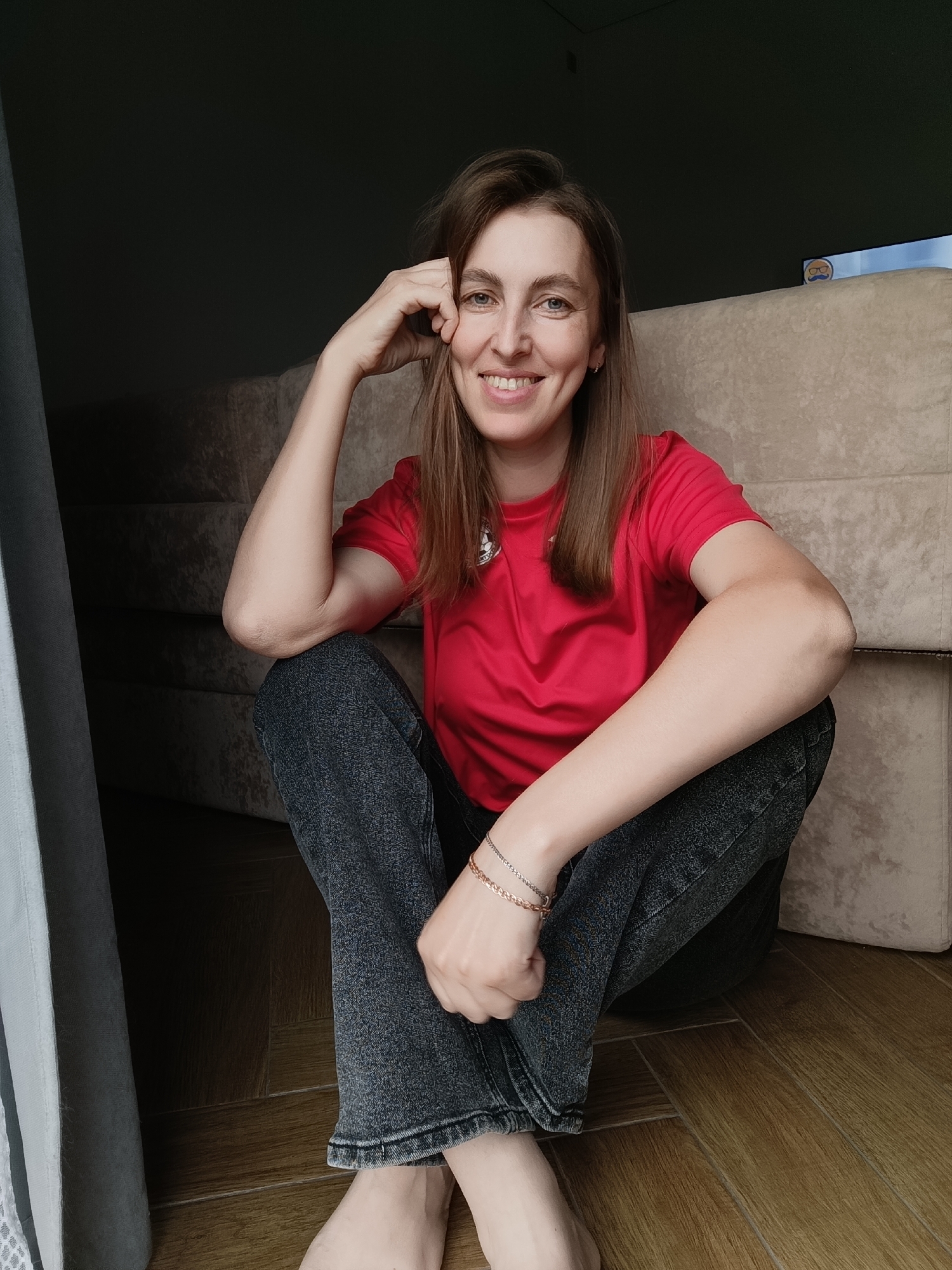 A young woman with light brown hair, wearing a red t-shirt and dark jeans, sits on a wooden floor with her legs crossed. She has her left arm bent, with her hand resting on her forehead, and she is smiling at the camera. She is wearing a bracelet on her right wrist. Behind her is a light-colored cushioned sofa, and to her left is a dark, shadowed area with a curtain. The scene appears to be indoors, possibly in a living room or common area. The lighting is soft, coming from the right side of the image. The overall impression is casual and relaxed. There is no explicit indication of the city of Chaikovsky, Russia, in the image itself.