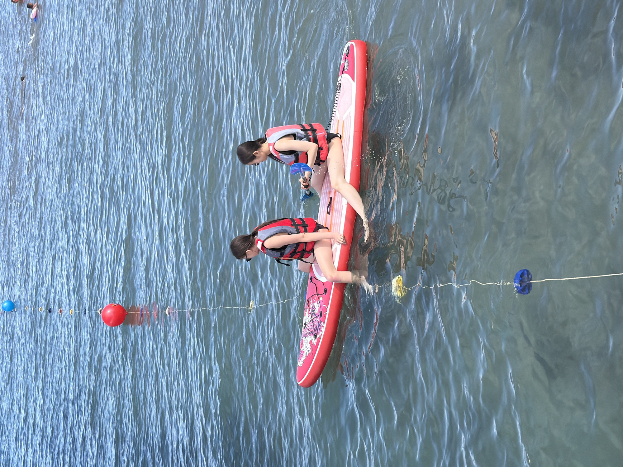 Two young women are paddleboarding on a calm body of water. They are both wearing red and grey life vests, suggesting a recreational or safety-conscious activity. The woman in front is kneeling on a red and white paddleboard, leaning forward and holding a blue paddle. The woman behind her is also kneeling on the same paddleboard, with her legs submerged in the water.
The paddleboard has a pink design on it, possibly a graphic. The water is a clear blue with ripples, indicating a peaceful and possibly sunny day. In the background, there is a rope with several colorful buoys, including a large red one and a blue one, which could mark a swimming area or a race course. The water appears to be shallow near the buoys. The overall mood seems relaxed and enjoyable, likely a summer day activity. There is no visible text in the image.
