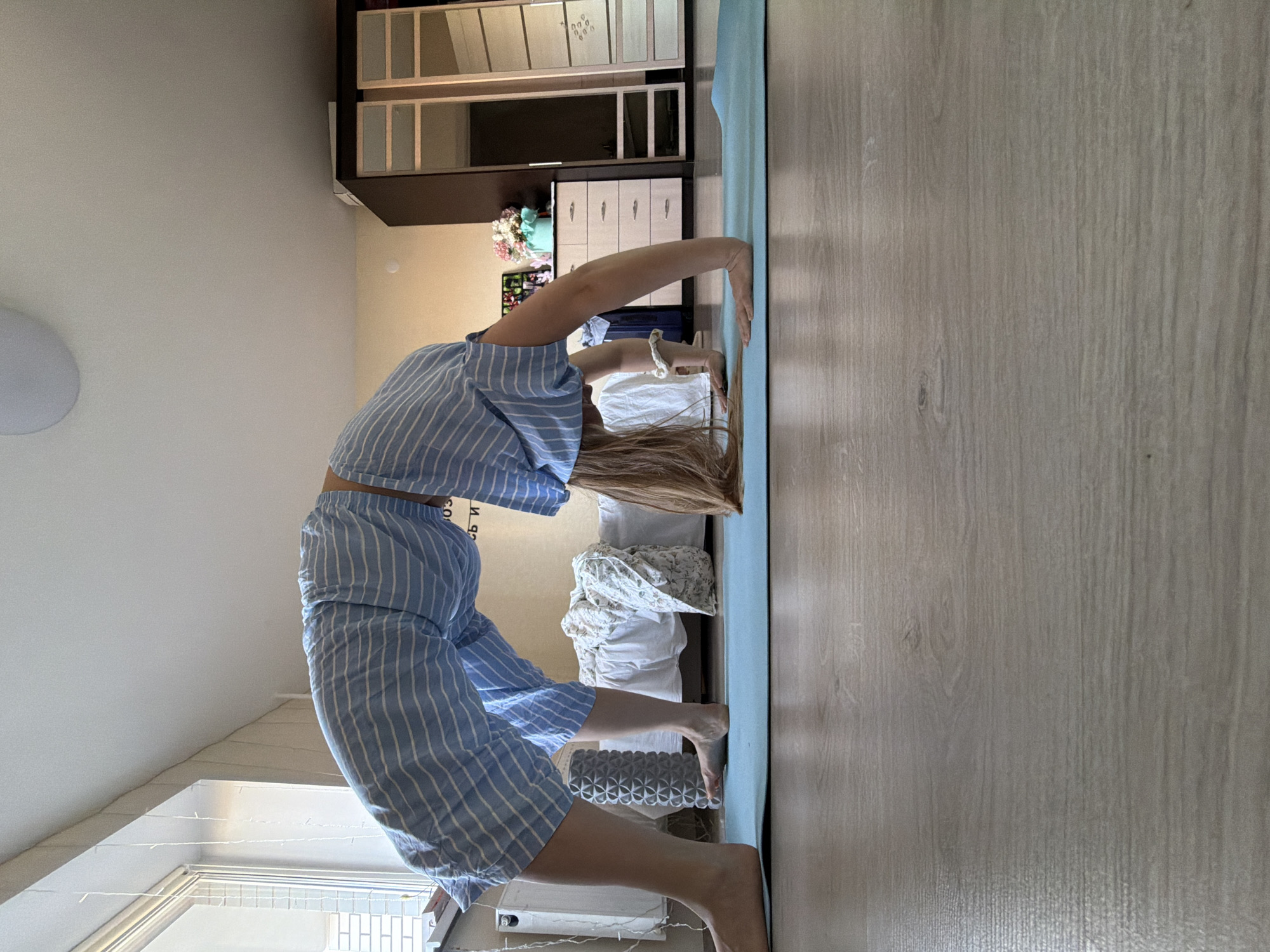 A young woman is performing a backbend yoga pose on a light blue yoga mat placed on a wood-patterned floor. She wears a striped blue and white pajama set. The background includes a wardrobe with mirrored and closed storage sections, a bed with rumpled white bedding, a white radiator, and a window with string lights. The location appears to be a room in a residential building. There is no evidence to identify the location is in Izhevsk, Russia.