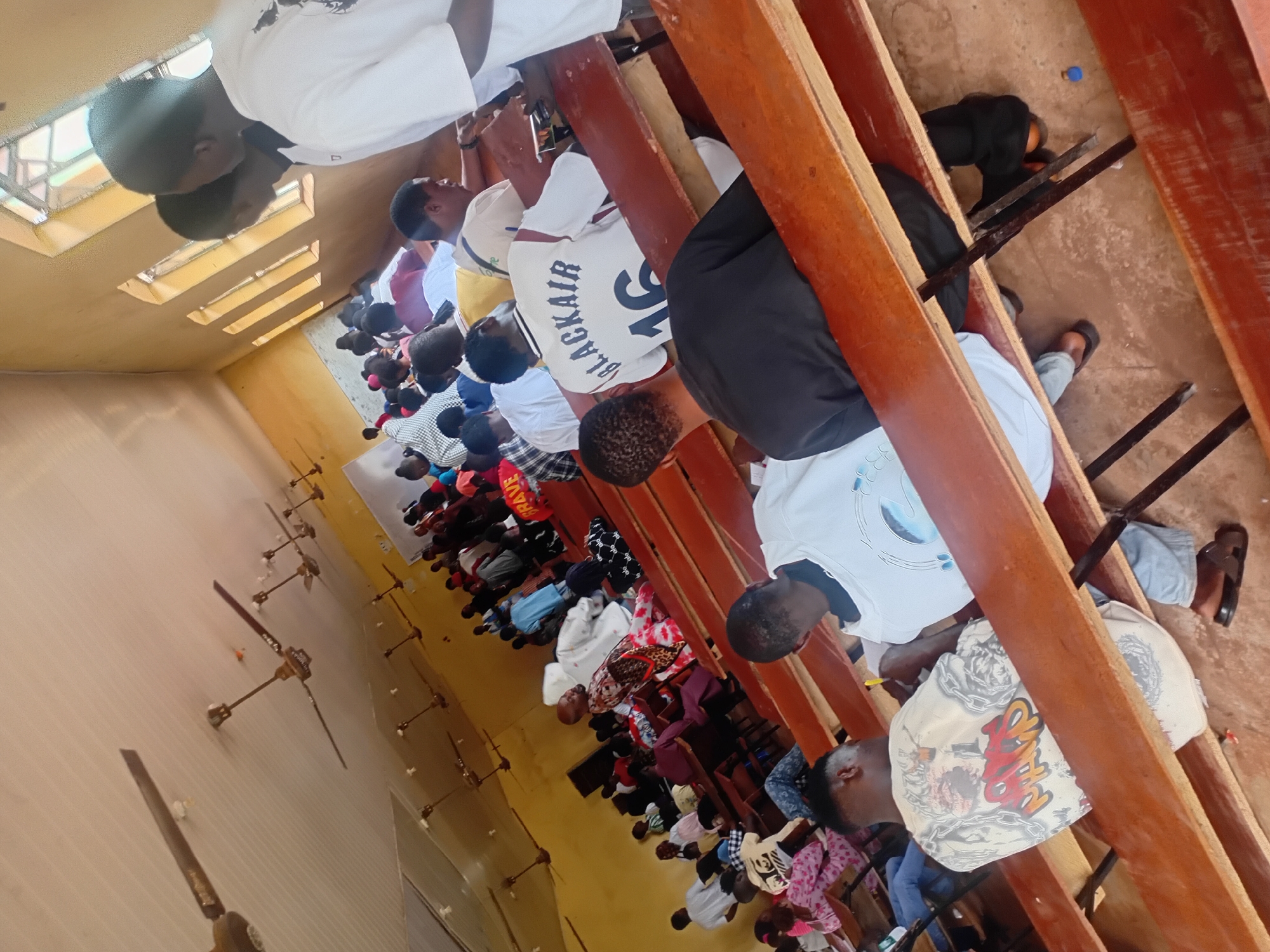 In Ogbogbo, Nigeria, a classroom of approximately 30 individuals, predominantly young men, are seated on wooden benches. The room has a textured, cream-colored ceiling with multiple ceiling fans, and the walls are painted a light yellow. Large, arched windows with segmented panes allow natural light to enter. The students are facing towards the front of the room, and some appear to be engaged in note-taking or listening. One student in the foreground is wearing a white t-shirt with a graphic design, and another student in the second row wears a white t-shirt with "BLACKAIR 16" printed on the back. The floor is made of packed earth or dirt.
