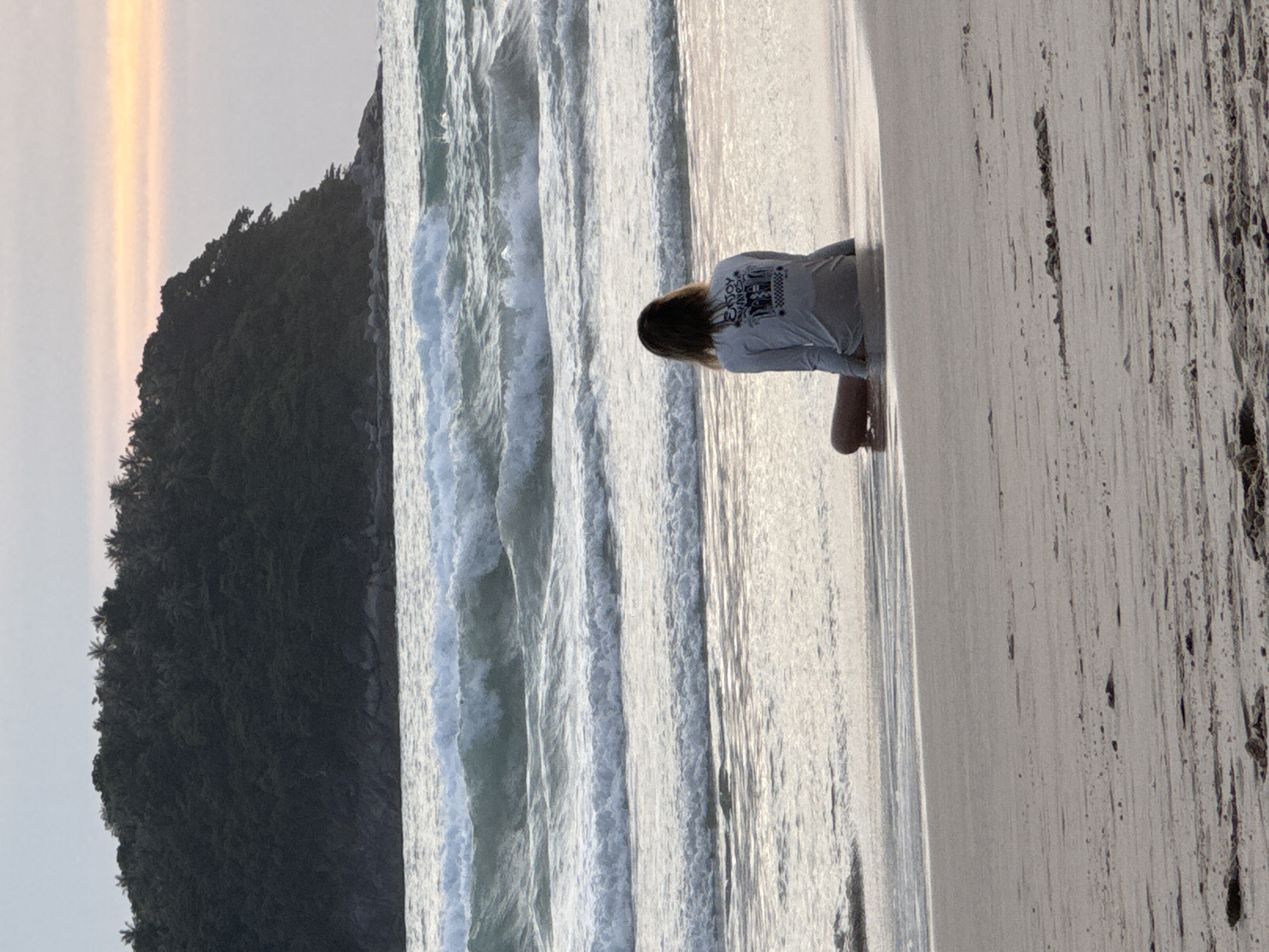 A person sits on a wet sandy beach, facing the ocean. The person is wearing a long-sleeved gray shirt with a graphic design on the back. Their legs are bent, with their feet partially submerged in the shallow water. The ocean has moderate waves, with white foam cresting. In the background, a tree-covered island juts out from the water. The sky above is overcast with a hint of orange and yellow near the horizon, suggesting either sunrise or sunset. The overall scene depicts a tranquil beach environment, likely in Karon, Thailand, given the tropical island vegetation and the style of the beach.