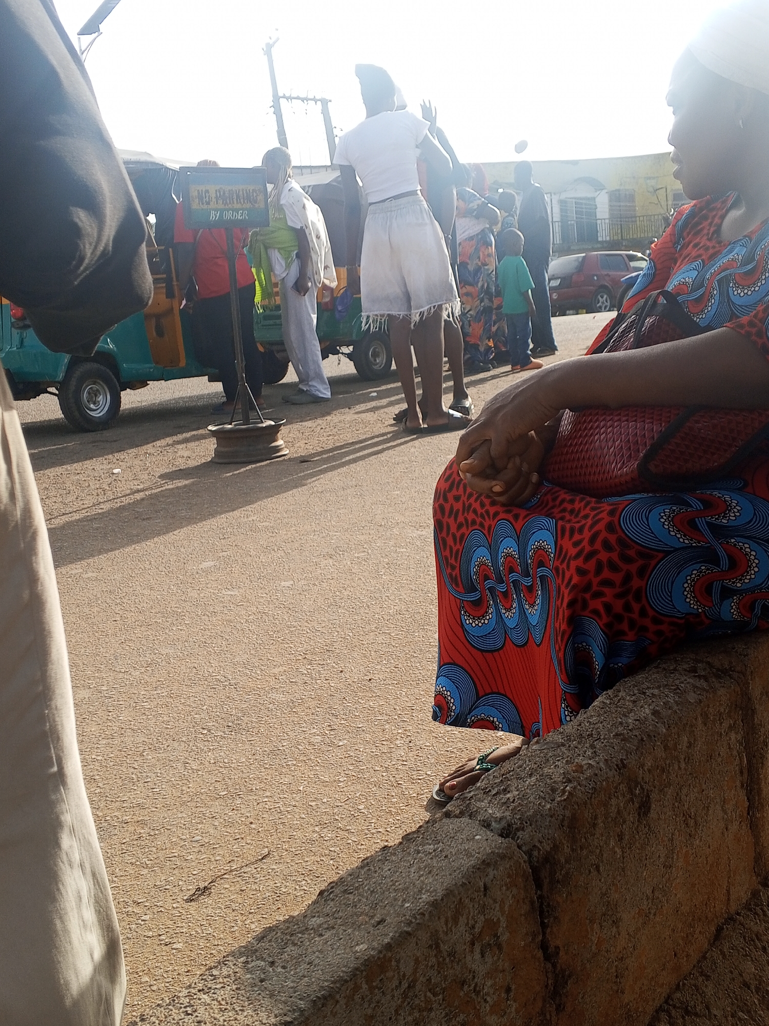 In Gwong, Nigeria, a woman in a red and blue patterned dress sits on a concrete barrier at the edge of a street. Her hands are clasped in her lap, and a purse rests on her lap. Behind her, a group of people are gathered near a green auto rickshaw. A "No Parking By Order" sign is visible next to the auto rickshaw. The street is paved and has a tan color. A red car is parked further down the street. The scene appears to be a busy public area.