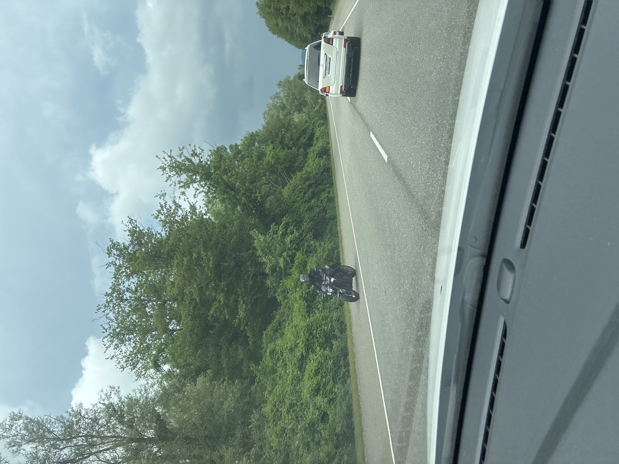An eye-level, medium shot captures a motorcycle rider on a highway lined with lush green trees and vegetation. The rider, wearing a dark helmet and jacket, is positioned in the left lane, appearing to travel towards the viewer. A white SUV is visible in the right lane, further down the road and also traveling away from the viewer. The highway is paved and has white lane markings. The sky above is overcast with a mix of grey and white clouds. The perspective suggests the image was taken from the dashboard of a vehicle, with the edge of the dashboard visible in the foreground. The location is identified as Arkhipo-Osipovka, Russia, implying a scenic, potentially coastal highway environment common in the region.