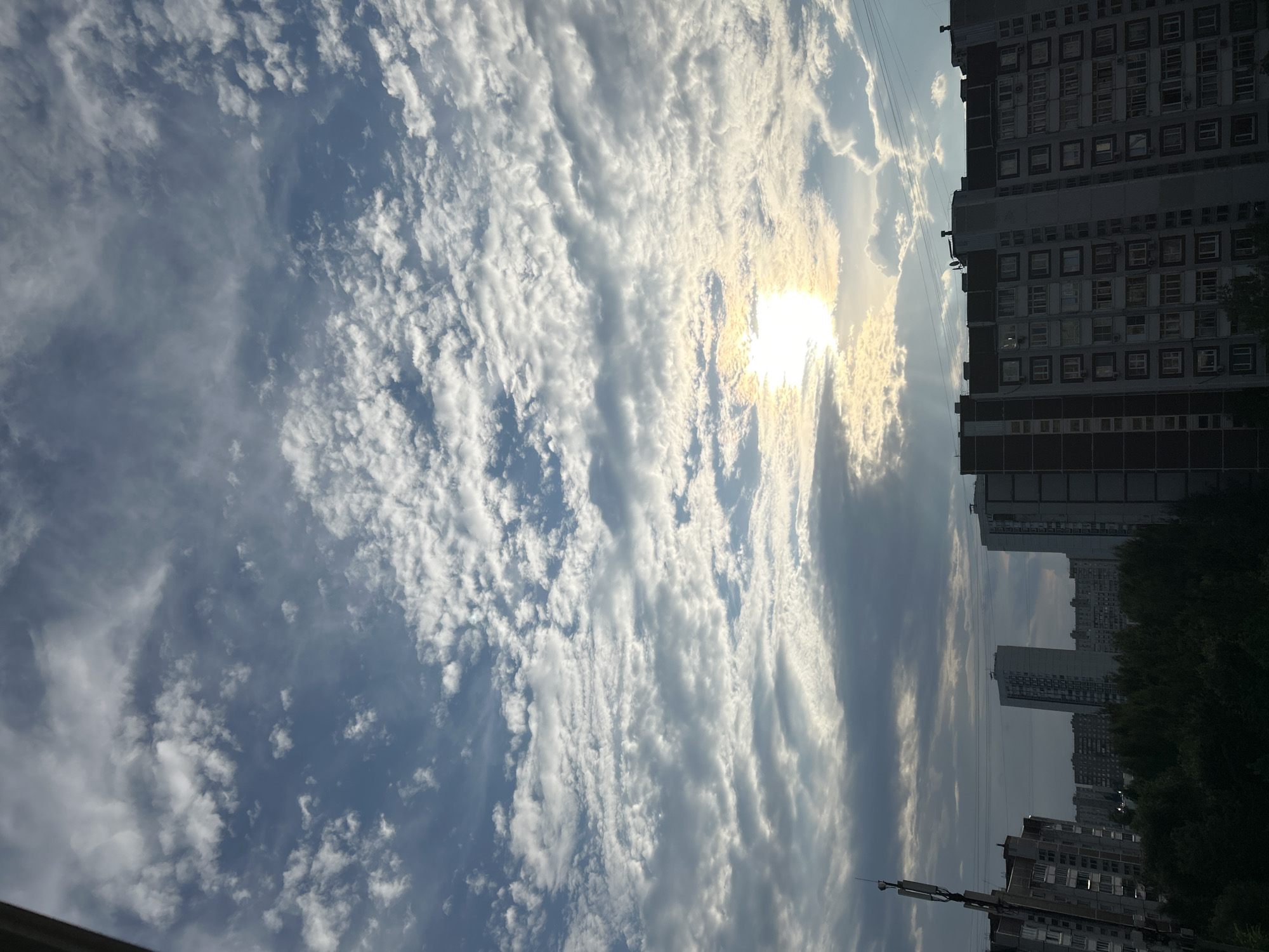 The image captures a view of the sky and buildings in Zelenograd, Russia, during what appears to be late afternoon or early evening. The sky is filled with a mix of cumulus and altocumulus clouds, with the sun partially obscured by them, creating a bright glow and visible sunbeams. Below the sky, a line of apartment buildings, characteristic of Soviet-era architecture, stretches across the frame. These buildings are multi-story, with numerous windows visible. Some buildings have antennas on their roofs. In the foreground, there are trees, indicating a green space within the urban environment. The overall impression is of a residential area in Zelenograd under a cloudy sky with the sun shining through.