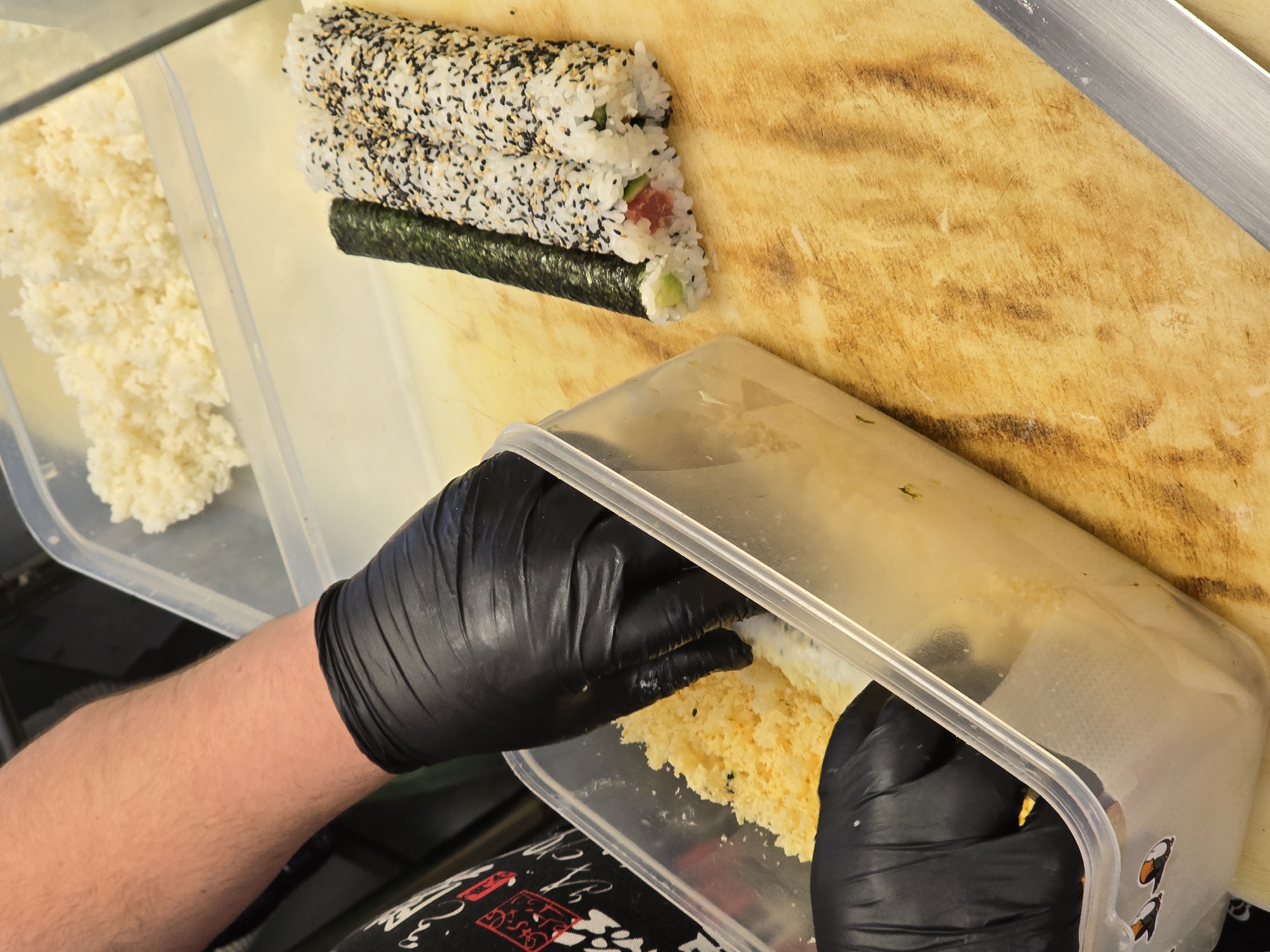 A close-up shows a person wearing black gloves preparing sushi in a kitchen setting, possibly in Burgas, Bulgaria. A sushi roll coated with black and white sesame seeds lies on a wooden cutting board. The person is reaching into a clear plastic container filled with yellow-tinted rice, possibly seasoned with egg. A separate container with white rice is visible on the left. A silver knife is partially visible on the right side of the cutting board. The person's attire includes a garment with a black and white pattern, with hints of red.