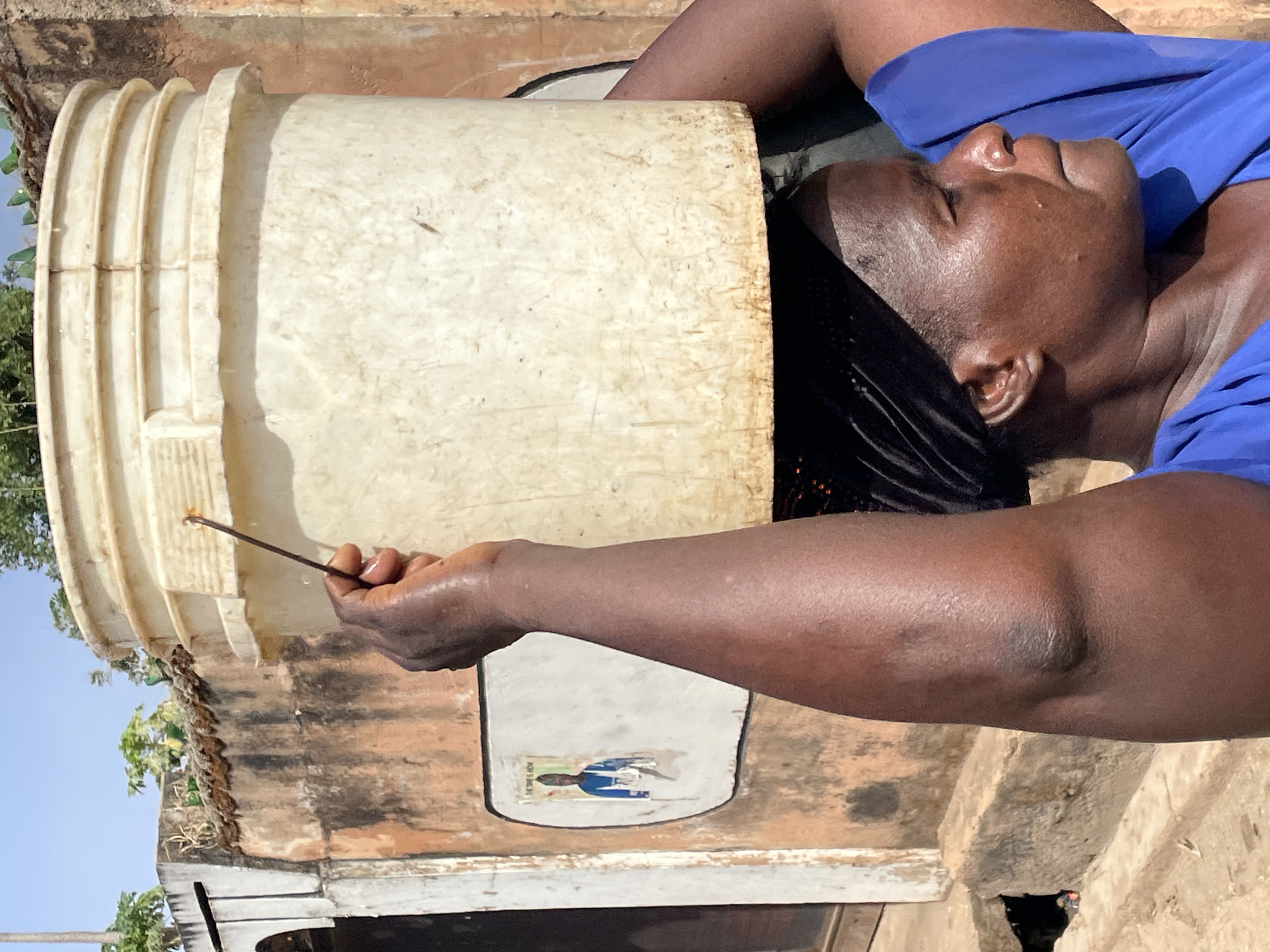 An adult person with dark skin, wearing a blue top and black braided hair, is depicted in an outdoor setting, likely in Jos, Nigeria. The individual is positioned with their head and upper body near or inside a large, off-white, aged plastic bucket that is lying horizontally. Their right arm is extended, holding a thin, dark stick with a light-colored, possibly amber or honey-like substance on its tip. The person's face is relaxed, eyes slightly downcast, suggesting concentration on the task at hand.
The background features a rustic, light brown wall made of concrete or mud bricks, showing signs of wear and age. A small, faded sticker or poster on the wall depicts a person in blue, with some barely legible text above it. Above the wall, green foliage and a clear, bright blue sky are visible, indicating sunny daytime weather. The scene appears to capture an activity related to the contents of the bucket, possibly collecting or inspecting honey or another sticky substance, given the material on the stick.