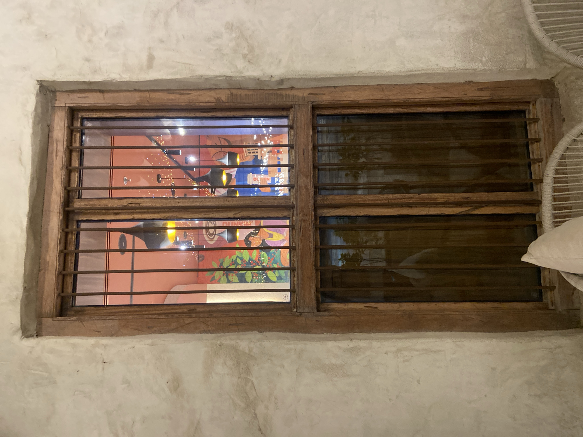 The image shows a rustic wooden-framed window with metal bars, set into a textured light-colored wall. The window is divided into two panes, with the upper pane reflecting the interior of a brightly lit establishment. Inside, the ceiling is painted orange, and several modern black pendant lights with yellow bulbs hang down. There are also strings of small fairy lights. A colorful mural or artwork is visible on the back wall, depicting a person in traditional clothing and foliage. The lower pane of the window is darker, showing reflections of the outdoor environment and a person or objects in front of the window, blurred by the glass and low light conditions. Part of a white woven chair and a light-colored cushion are visible in the foreground at the bottom of the image. The overall impression is of a dimly lit interior viewed through a securely barred window, suggesting a cozy or perhaps somewhat secluded atmosphere, possibly in a cafe or restaurant in Deira, United Arab Emirates.