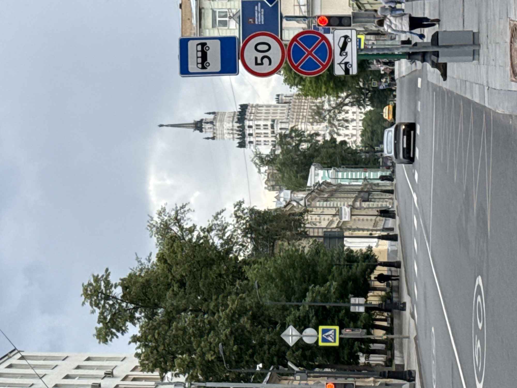 The image depicts a street scene in Moscow, Russia, likely on a cloudy day. A distinctive Stalinist skyscraper with a spire dominates the background skyline. In the foreground, a two-lane asphalt road runs through the center of the frame, with white lane markings visible. Several traffic signs are prominently displayed on the right side of the road, including a bus stop sign, a 50 km/h speed limit sign, a no stopping sign, and a towing sign. A traffic light is also visible, showing red.
On the sidewalk to the right, several pedestrians are walking, including a person in a white vest and another in a gray jacket. A trash receptacle is also present. A dark-colored car is driving in the road, and a yellow taxi is seen further down the street.
On the left side of the road, trees with dense green foliage line the sidewalk. A pedestrian crossing sign with a yellow diamond shape is mounted on a pole. A traffic signal is also on this side, showing red. Buildings with multiple windows and some architectural detail are visible along the street. The overall impression is of a typical urban environment with busy street traffic and pedestrian activity.
