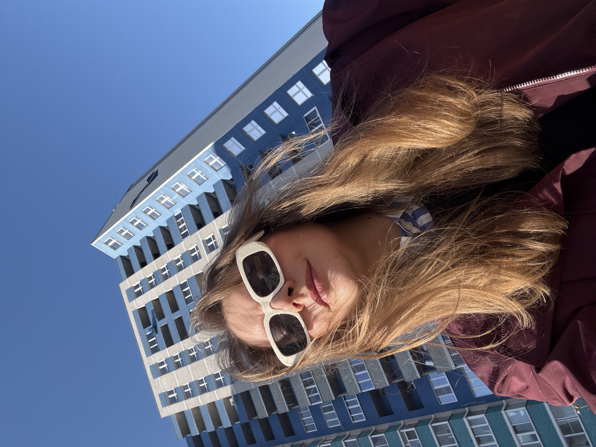 A low-angle shot shows a woman with long blonde hair and white-framed sunglasses, presumably taking a selfie. She is wearing a maroon jacket. Behind her is a tall building with a blue and gray exterior and numerous windows. The sky is a clear, bright blue. The building appears to be located in Izhevsk, Russia.