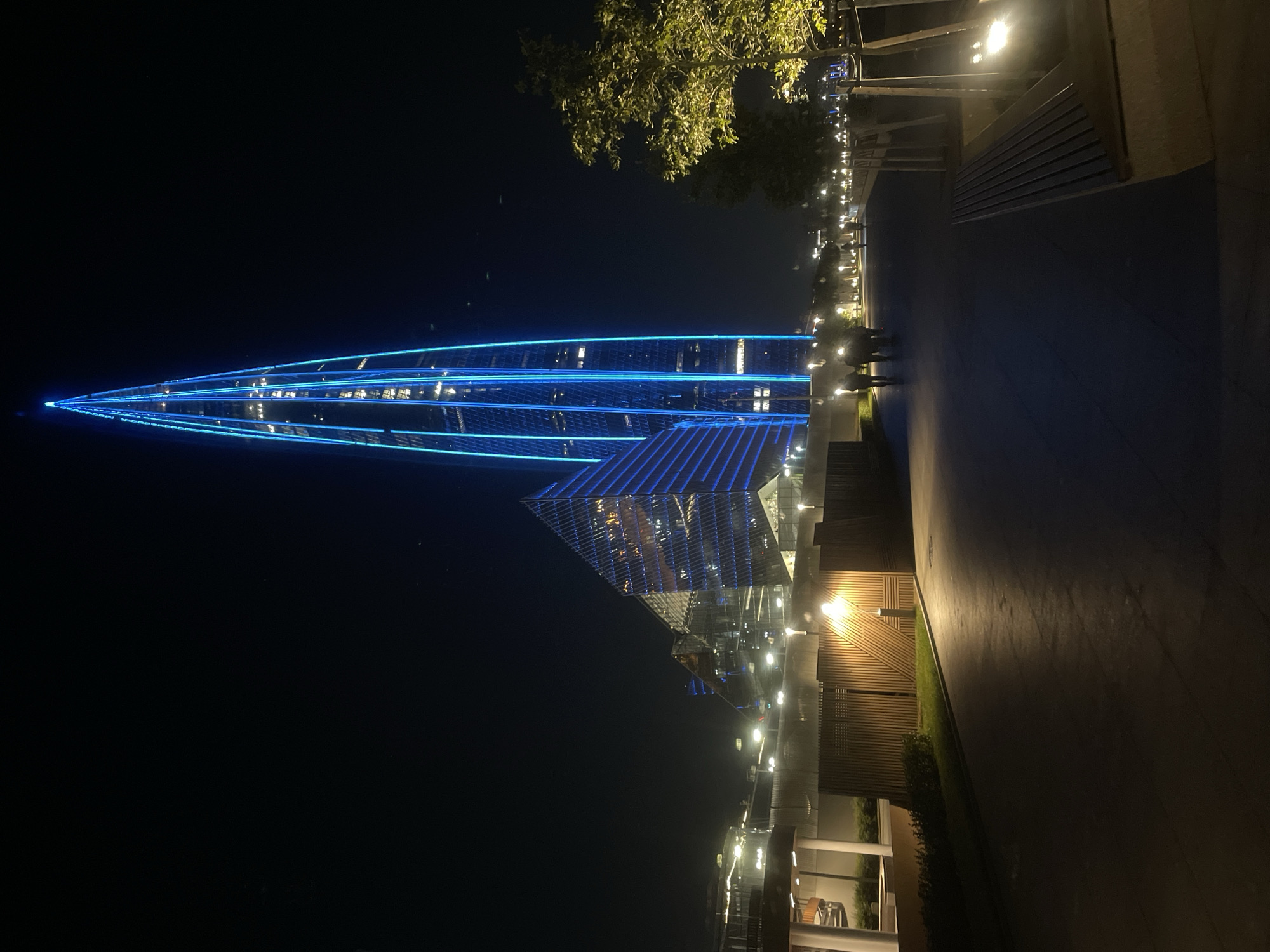 The image depicts the Lakhta Center in Saint Petersburg, Russia at night. The prominent feature is the skyscraper, illuminated with bright blue lights that accentuate its spire. Adjacent to the skyscraper, another building with a faceted glass facade also displays blue lighting. In the foreground, a paved walkway stretches into the distance, bordered by wooden fencing and lampposts that cast warm light on the scene. Several people are visible walking along the path, silhouetted against the illuminated cityscape. A tree with vibrant green leaves stands to the right of the walkway, also lit by a nearby lamp. The overall atmosphere is one of modern urban development set against the darkness of night.
