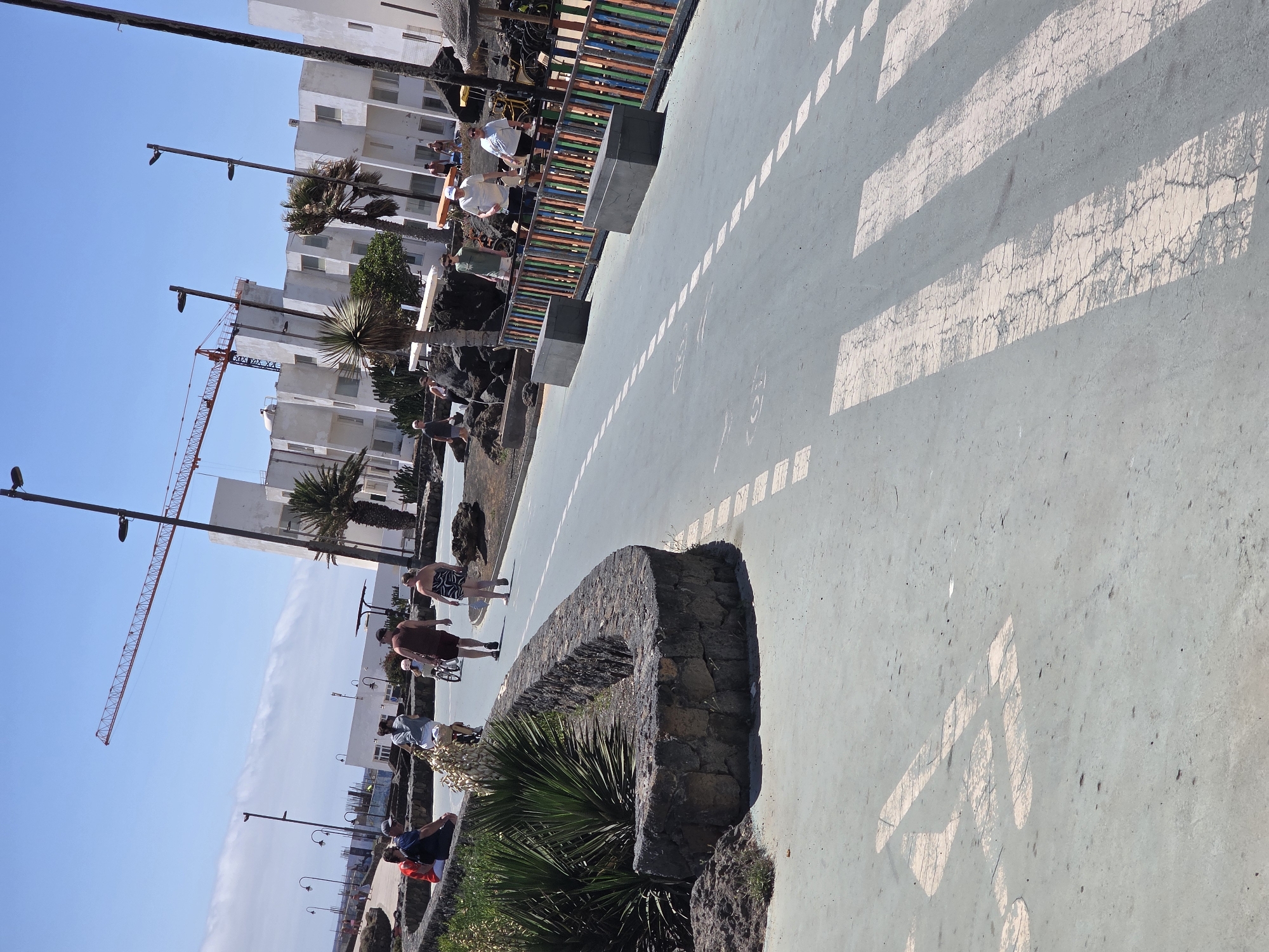 A wide shot captures a sunny day scene in Costa Teguise, Spain, with a focus on a pedestrian walkway. In the foreground, a light blue asphalt surface is marked with white pedestrian crossing lines and a bicycle symbol. To the left, a low, curved stone wall borders a patch of green vegetation and dark volcanic rock. Further back, a paved walkway runs parallel to a colorful fence made of vertical wooden slats in red, orange, yellow, green, and blue.
Several people are visible on the walkway. On the left side, near the stone wall, a group of three individuals, two seated and one standing, are dressed casually. Further along the walkway, a person is riding a bicycle, followed by another person walking. A small child is seated on a bicycle behind them. To the right of the walkway, more individuals are seen strolling, some in swimwear.
In the background, modern white buildings with multiple windows stand under a clear blue sky. A prominent red construction crane is visible against the sky, indicating ongoing development. Palm trees and other tropical vegetation are scattered throughout the scene. The overall environment suggests a resort or coastal town setting.