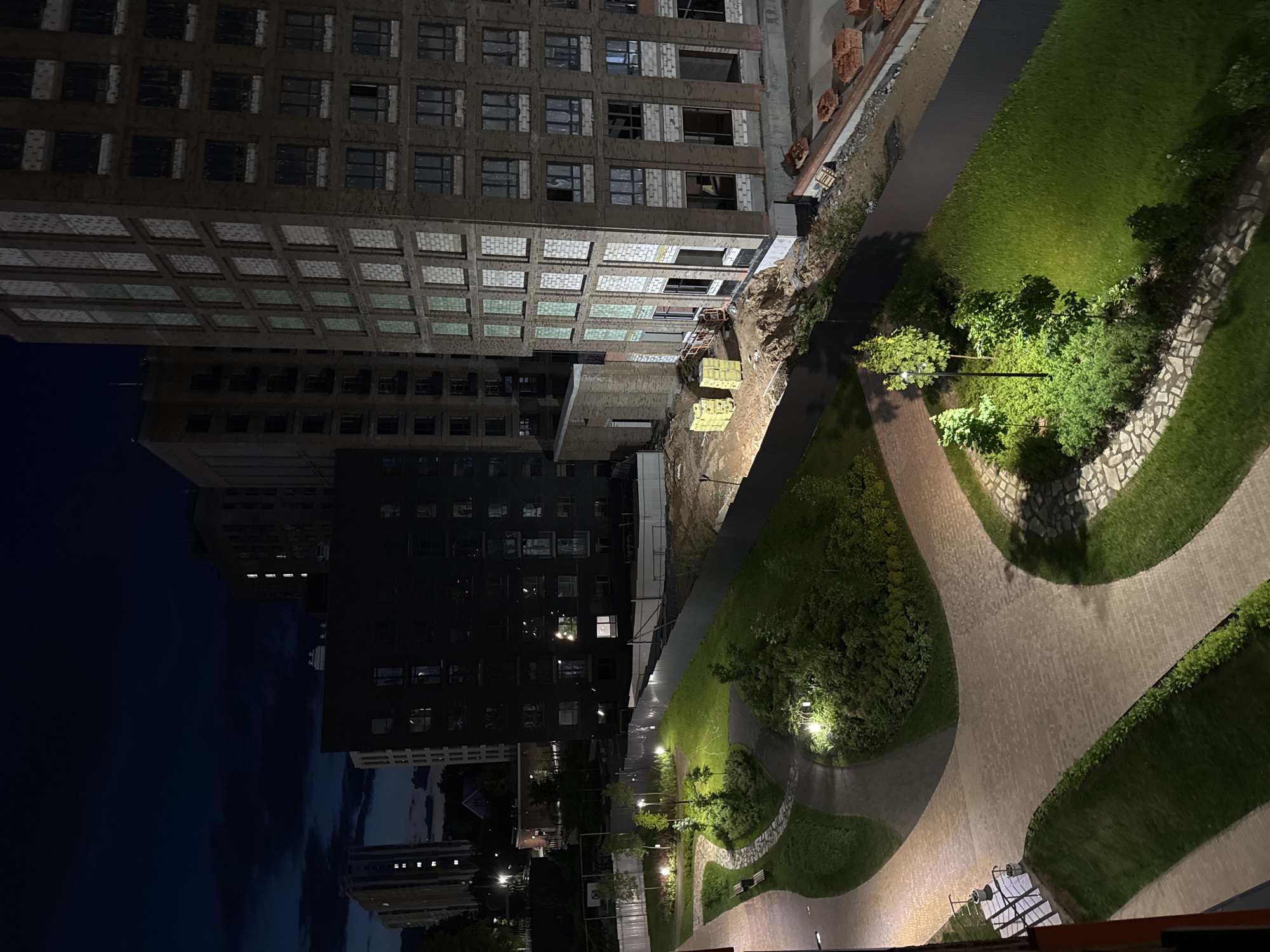 This media file presents a nighttime urban scene, taken from an elevated perspective, likely a balcony or higher floor, looking down and across an area in Izhevsk, Russia. The image is oriented vertically, with buildings appearing to lie on their sides; however, describing it as if rotated 90 degrees clockwise shows a typical upright urban landscape.
In the foreground, a well-lit park or courtyard area features curving paved pathways, neatly trimmed grass sections, and several small trees and bushes. The paths are illuminated by multiple spherical streetlights, casting a warm glow on the greenery. Some path edges are bordered with decorative stonework.
Immediately beyond the park, a transition zone shows elements of an active construction site with disturbed earth, a temporary fence, and stacked building materials such as pallets of bricks or concrete blocks.
The midground is dominated by multi-story residential buildings. On the right, one building appears to be actively under construction, with exposed brick and concrete elements, unfinished window openings, and some scaffolding visible. Adjacent to it, another building, possibly part of the same complex, looks more complete, with numerous windows. On the left, a tall, dark residential building stands prominently, its many illuminated windows suggesting occupied apartments.
In the background, the dark night sky shows faint cloud formations. More distant, dark urban structures with scattered lights are visible against the horizon. No people or legible text are visible in the image. The scene conveys a sense of ongoing urban development and residential life after dark.