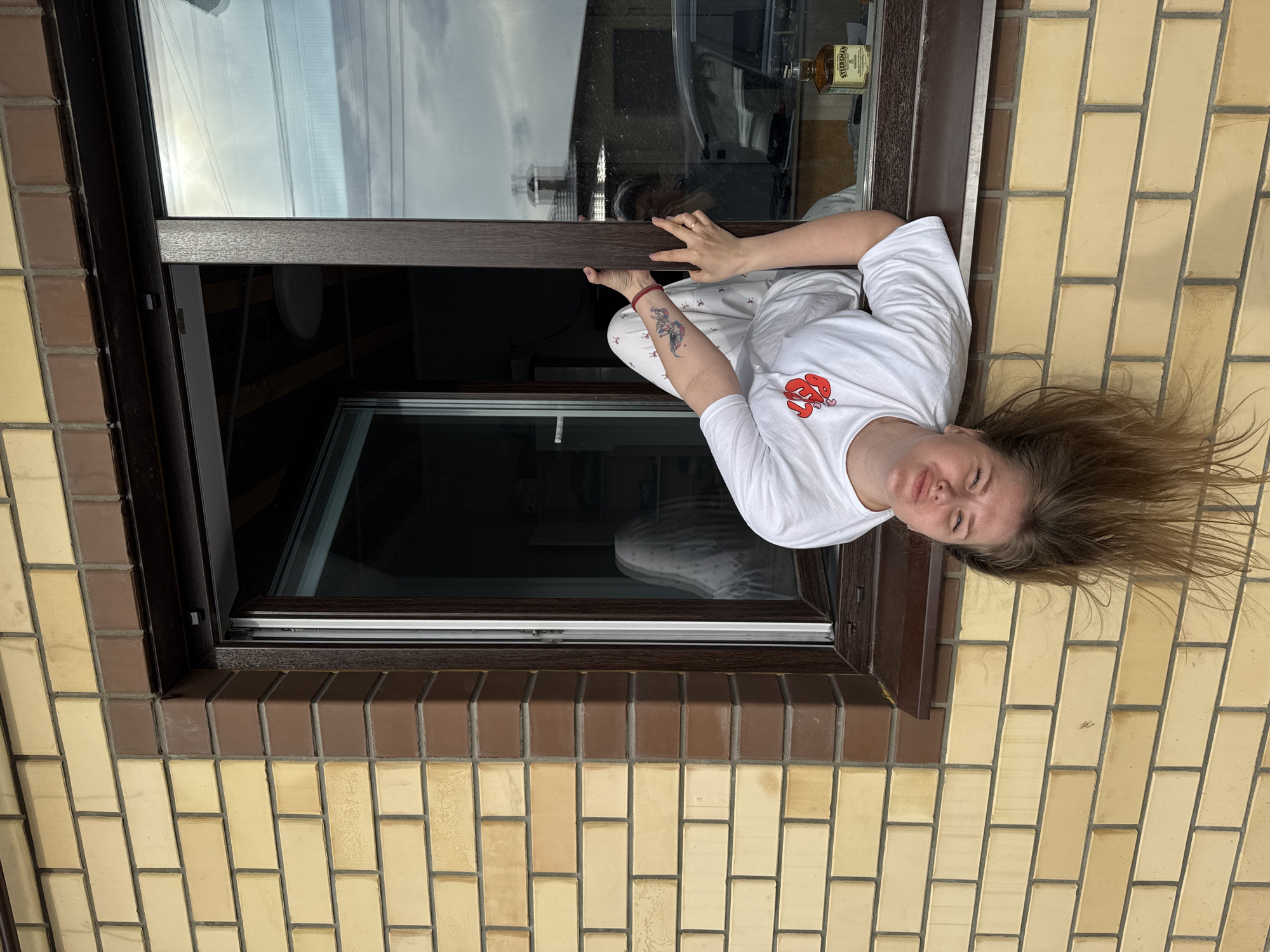 A woman is partially visible in a window of a building constructed of yellow brick with brown trim. The window is open, and the woman is leaning partway out. She is wearing a white t-shirt and pajamas and has a tattoo on her left forearm. A bottle of liquor is visible in the background through the window. The location is Gornyi (Dvurechenskoe S/P), Russia.