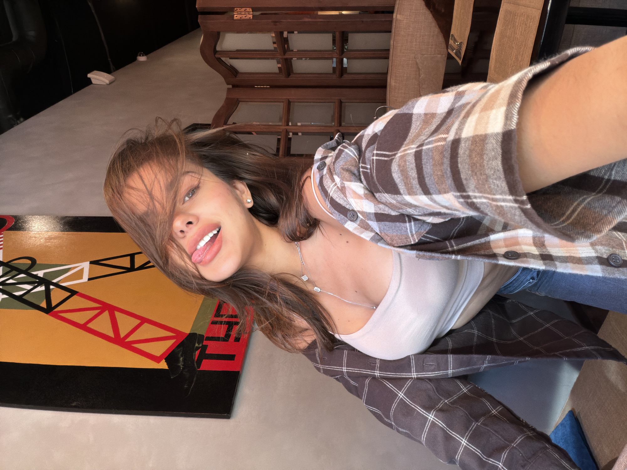 A selfie shows a young woman with long brown hair, wearing a white tank top under a brown and white plaid shirt, sticking her tongue out. She is wearing a silver necklace. A painting with black, yellow, white, and red geometric shapes is visible on the left. In the background, a wooden structure can be seen. The location is Krasnoyarsk, Russia.