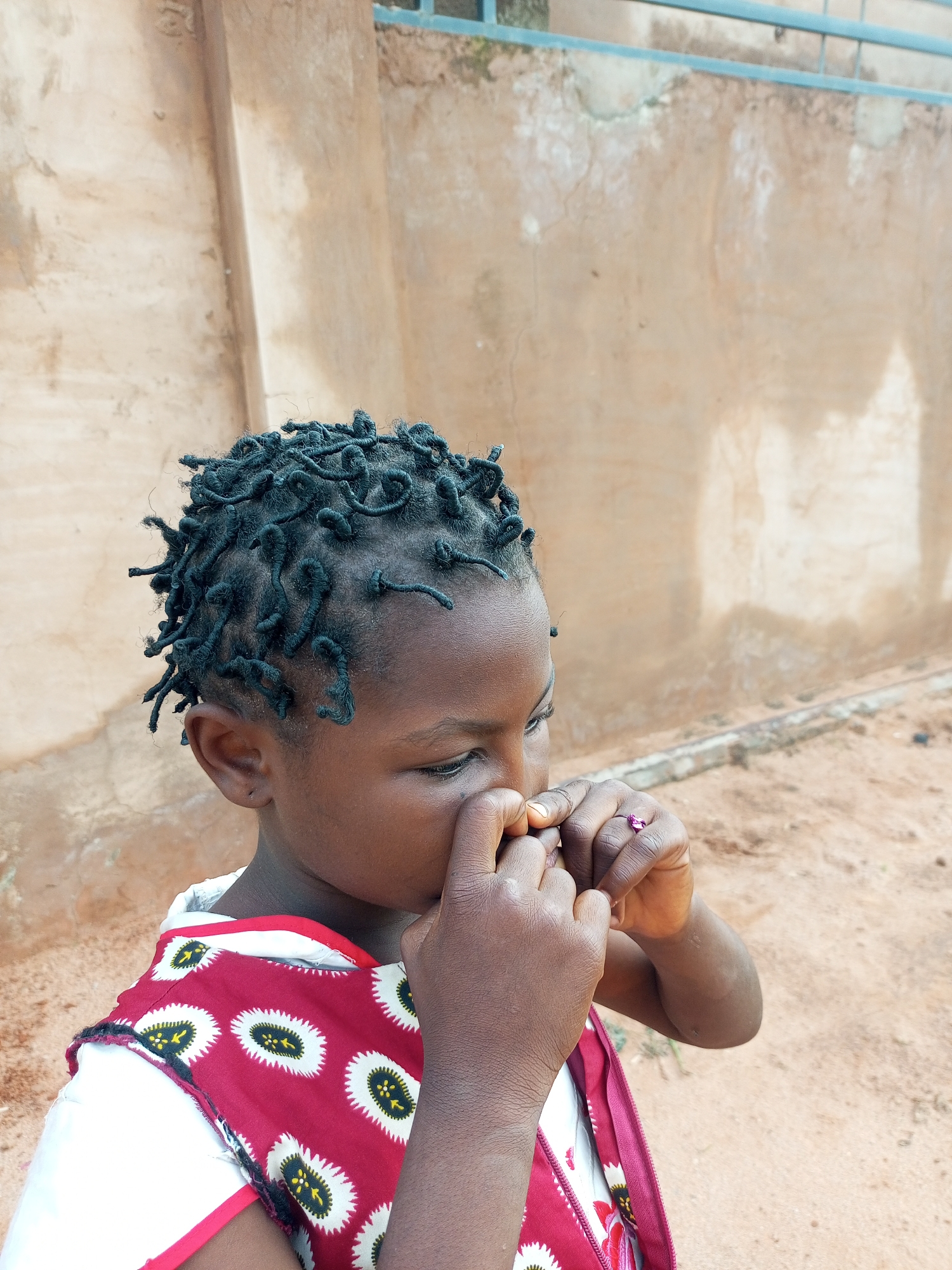 In Tempure, Nigeria, a young girl with dark skin and intricate braided hair is captured in a profile view, facing towards the right of the frame. She is wearing a white garment with a red overlay that features a repeating pattern of white oval shapes with black and yellow markings. Her hands are brought up to her face, with her fingers touching her nose. On her right ring finger, a small pink ring is visible. The background consists of a textured, light brown wall, possibly made of plaster or mud brick, with a horizontal metal railing running along the top. The ground in front of the wall appears to be sandy or dusty.
