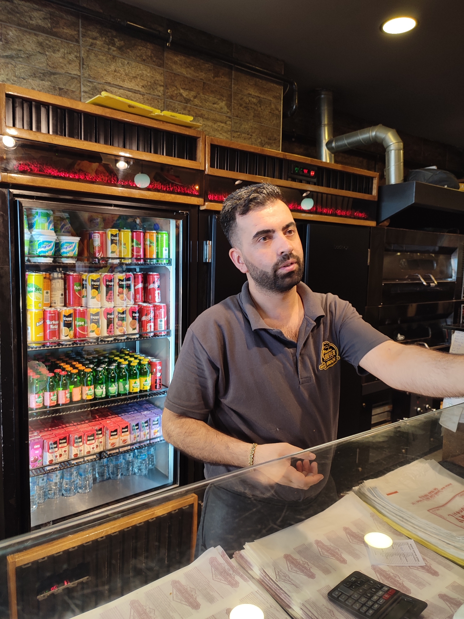 A man with dark hair and a beard, wearing a grey polo shirt with a yellow logo, stands behind a glass counter in what appears to be a food establishment. He is looking to his right and holding a calculator with his left hand, while his right arm is extended slightly. Behind him, a glass-fronted refrigerator is filled with various drinks, including what look like yogurt containers, canned beverages, and bottled juices. To the right of the refrigerator, a black oven is visible. The man's shirt has a logo that reads "SÖRFEYE BÖREKCISi." The environment suggests a retail setting, likely a bakery or a fast-food restaurant, possibly located in Kadıköy, Turkey, given the presence of Turkish language on some of the products. The counter is cluttered with papers and a calculator.