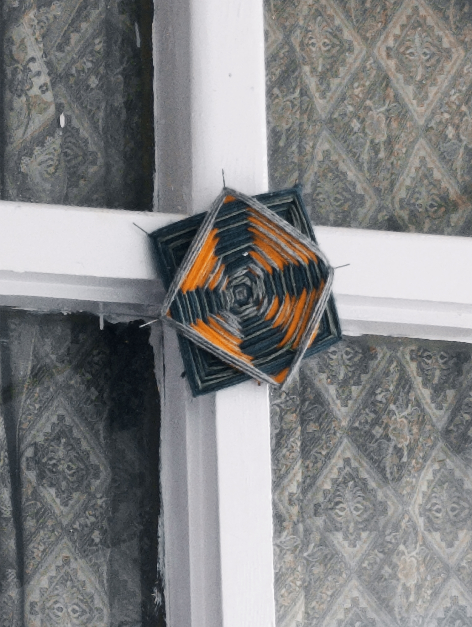 A hand-woven, diamond-shaped eye of God decoration, made with dark blue and orange yarn, is hanging on a white window frame in Vashisht, India. The decoration is centrally positioned, overlapping two panes of the window. Behind the window are sheer curtains with a damask pattern in muted tones of gray and beige. The window frame is painted white and appears to be part of a building, with a section of the wall visible to the right of the frame. The scene is lit by ambient light, creating a slightly soft focus effect. There are no people present in the image.