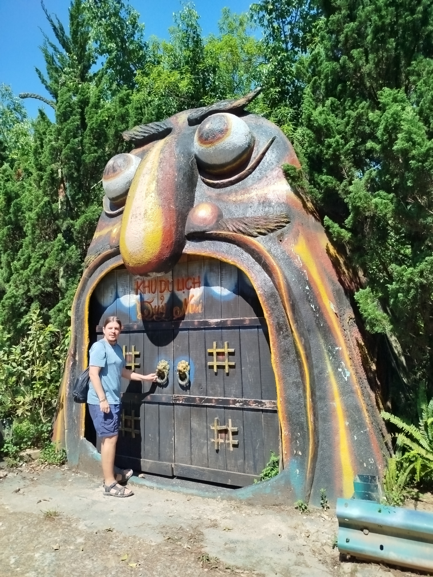 A man stands smiling in front of a large, whimsical face-like structure, serving as an entrance, in Păng Tiêng 1, Vietnam. The structure is designed to resemble a monstrous or troll-like face with large eyes, an elongated nose, and a wide-open mouth forming an archway for two dark wooden doors. The surface of the structure is textured and painted in earthy tones of brown, orange, yellow, and black.
The wooden doors feature decorative square metal grates and ornate golden lion's head door knockers. Above the doors, Vietnamese text reads "KHU DU LỊCH" (Tourist Area), with additional, partially obscured text below it.
The man is dressed in a light blue t-shirt, blue shorts, and sandals, carrying a black backpack. He is positioned to the left of the doors, with his right hand resting near one of the lion-head knockers, indicating he is posing for a photo. His expression is one of calm enjoyment.
The scene is outdoors, bathed in bright sunlight under a clear blue sky, suggesting a clear day. The background is filled with lush green foliage and tall trees, some appearing coniferous. The ground is a dirt path with scattered leaves and sparse vegetation. A teal-colored metal object, possibly part of a barrier, is visible in the lower right foreground.