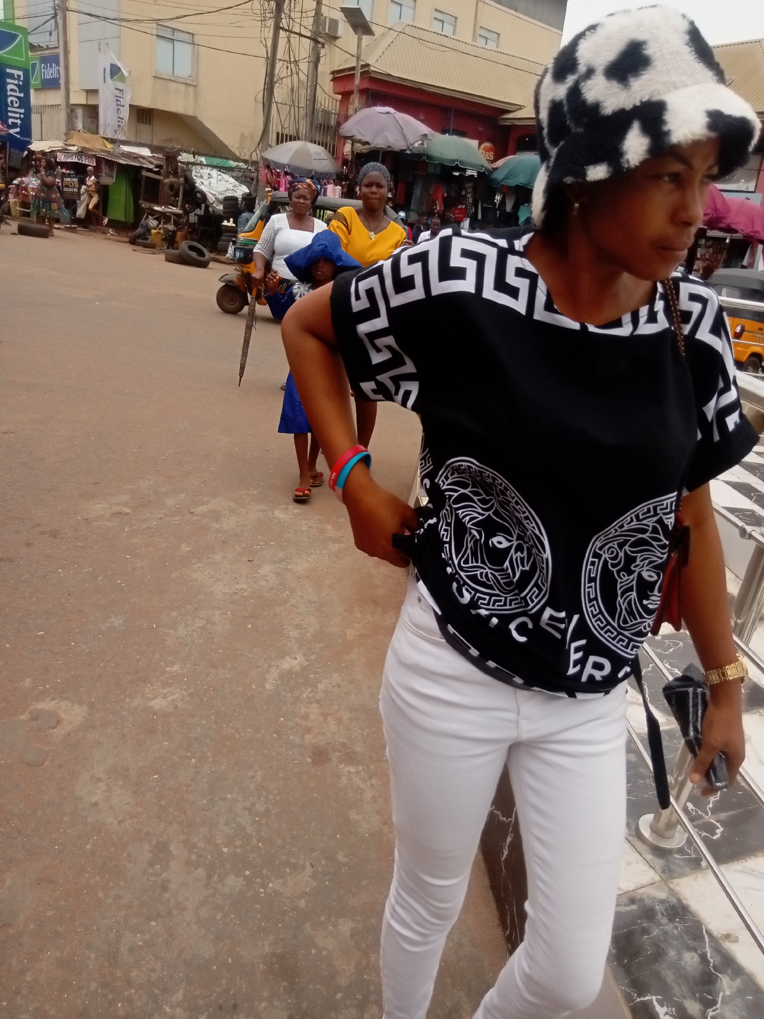 In Nkpor, Nigeria, a woman in a black and white patterned shirt, white pants, and a cow-print hat stands on a sidewalk. In the background, two women and a child walk down a bustling street with small shops and various vehicles. A "Fidelity" bank sign is visible in the upper left corner.