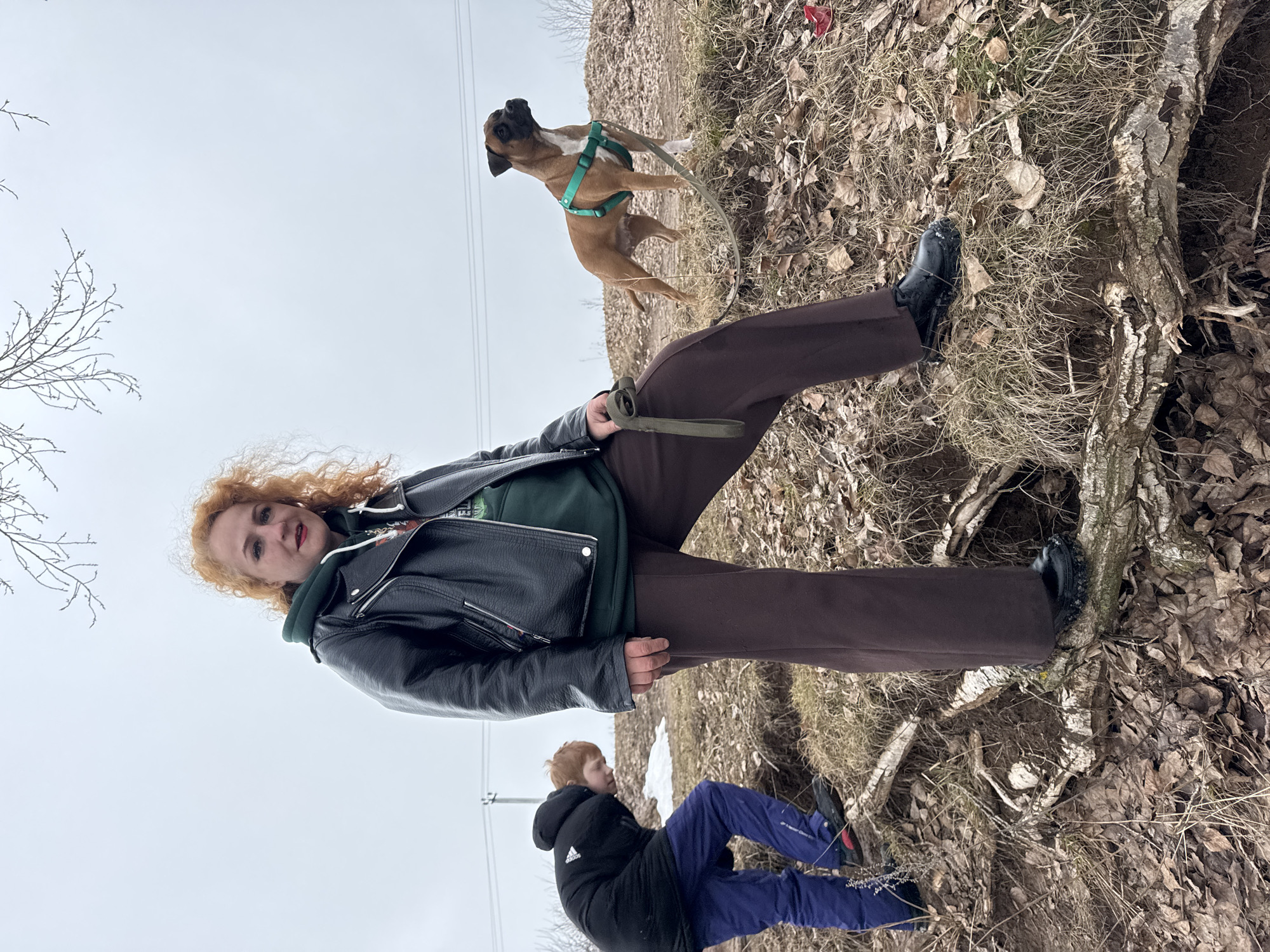 The image captures a scene from Chaikovsky, Russia. A woman with curly red hair, wearing a black leather jacket over a green hooded sweatshirt and brown pants, holds a dog leash. The dog, a tan Boxer with a white chest and green harness, is in mid-air, suggesting it is jumping or playing. A child in a black jacket and blue pants lies on the ground. The environment consists of dry leaves, grass, and tree roots, indicative of a natural area or park. Overcast skies suggest dull lighting.