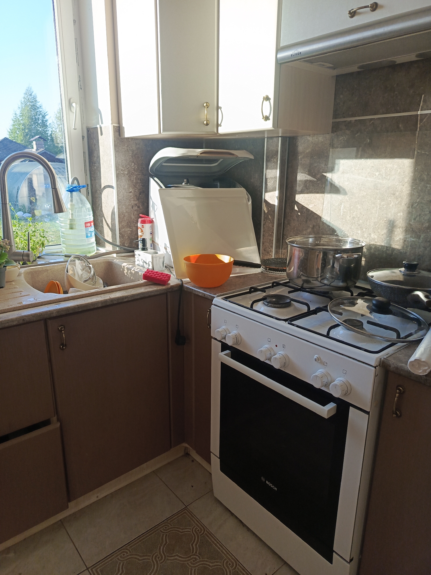 The image shows a kitchen scene in an indoor setting. A sink is situated near a window that reveals foliage and a building. On the counter surrounding the sink are several items, including a large jug of water and various dishes. To the right of the sink, a white gas stove is prominently featured, with pots and pans placed on top of it. The backdrop consists of kitchen cabinets and a tiled backsplash. The floor is tiled with a patterned design.