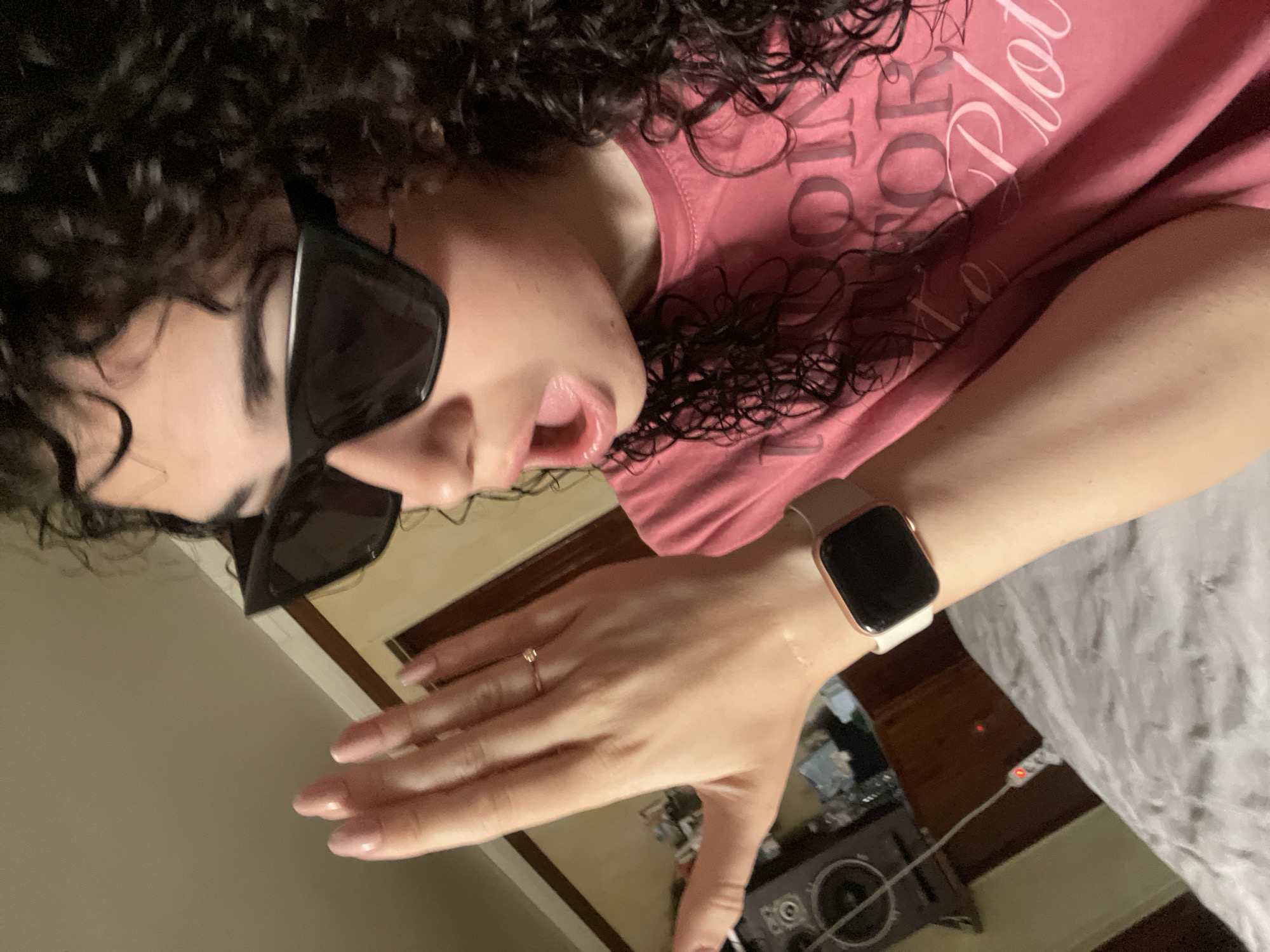 A woman with curly hair and sunglasses is wearing a pink t-shirt and an Apple Watch. She has a ring on her finger. The background includes a bed and a speaker. The image does not contain any information that would allow one to determine the location in Sofia, Bulgaria.
