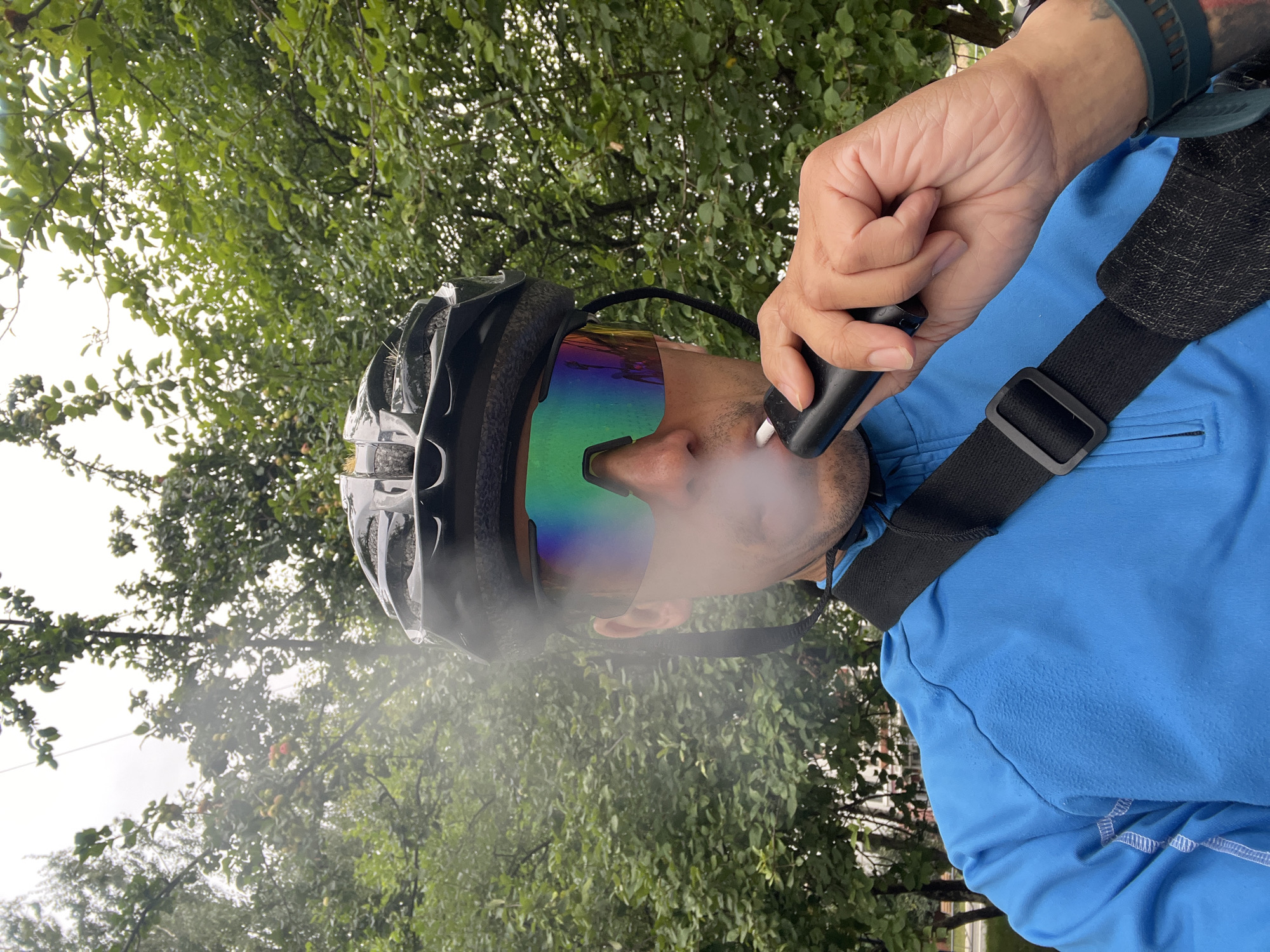 A close-up, selfie-style, medium shot shows a person wearing a black bicycle helmet and mirrored sports glasses with a green and blue iridescent tint. The person is wearing a bright blue long-sleeved athletic shirt and a black strap across their chest. They are holding a black electronic cigarette device to their mouth, exhaling a visible plume of vapor. The background consists of lush green foliage from trees. The image appears to be taken outdoors in a park-like setting, possibly in the city of Chaikovsky, Russia, given the presence of deciduous trees and what might be a suburban or park environment.