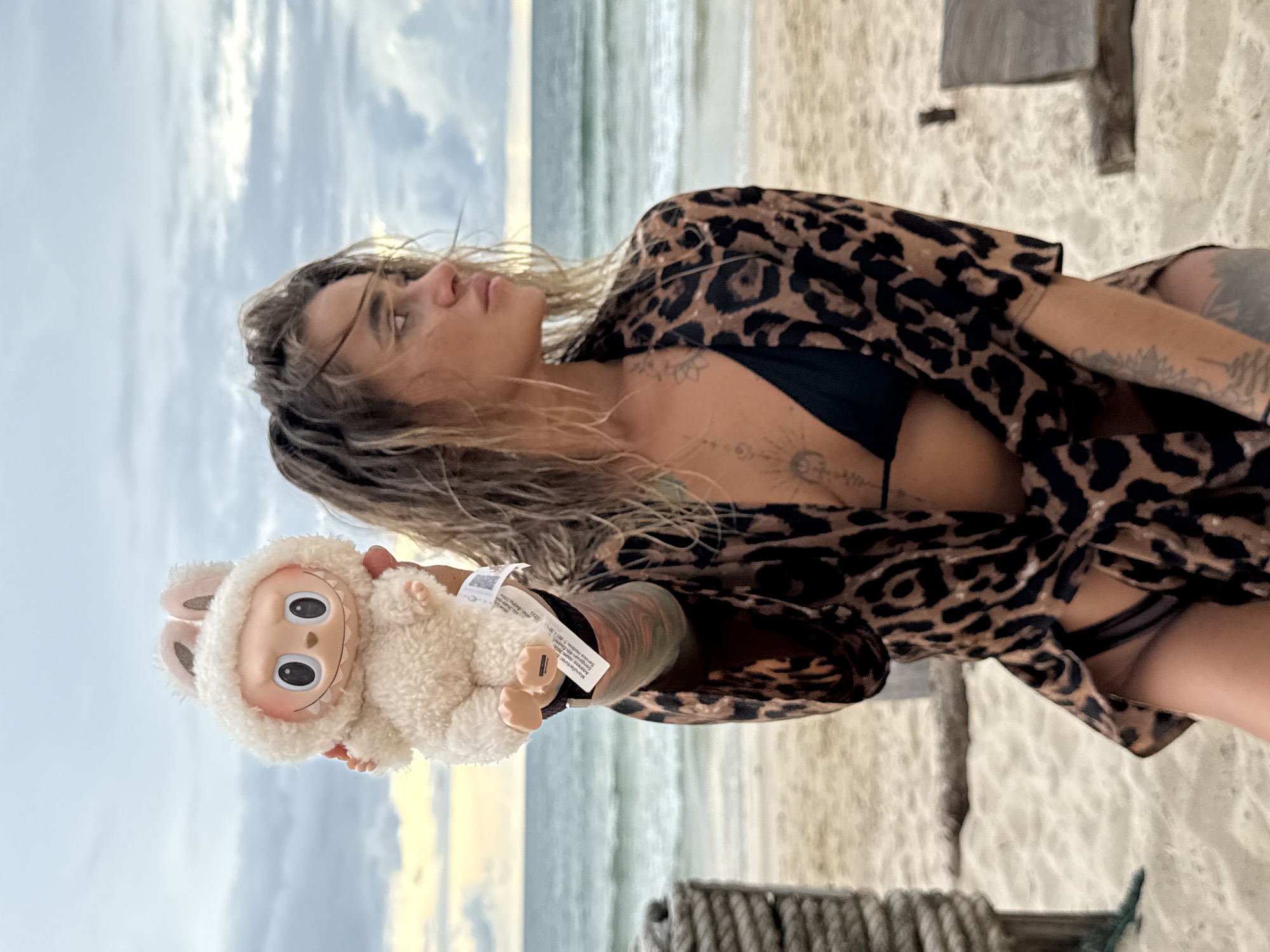 A woman with damp, wavy blonde hair, wearing a black bikini top and an open leopard print cover-up, stands on a sandy beach in Choeng Thale, Thailand. She is holding a small, fluffy white toy with bunny ears and a pink nose up with her left hand. The woman has tattoos visible on her arms, chest, and torso. The background features the ocean with gentle waves under a cloudy sky. The lighting suggests it is either dawn or dusk.