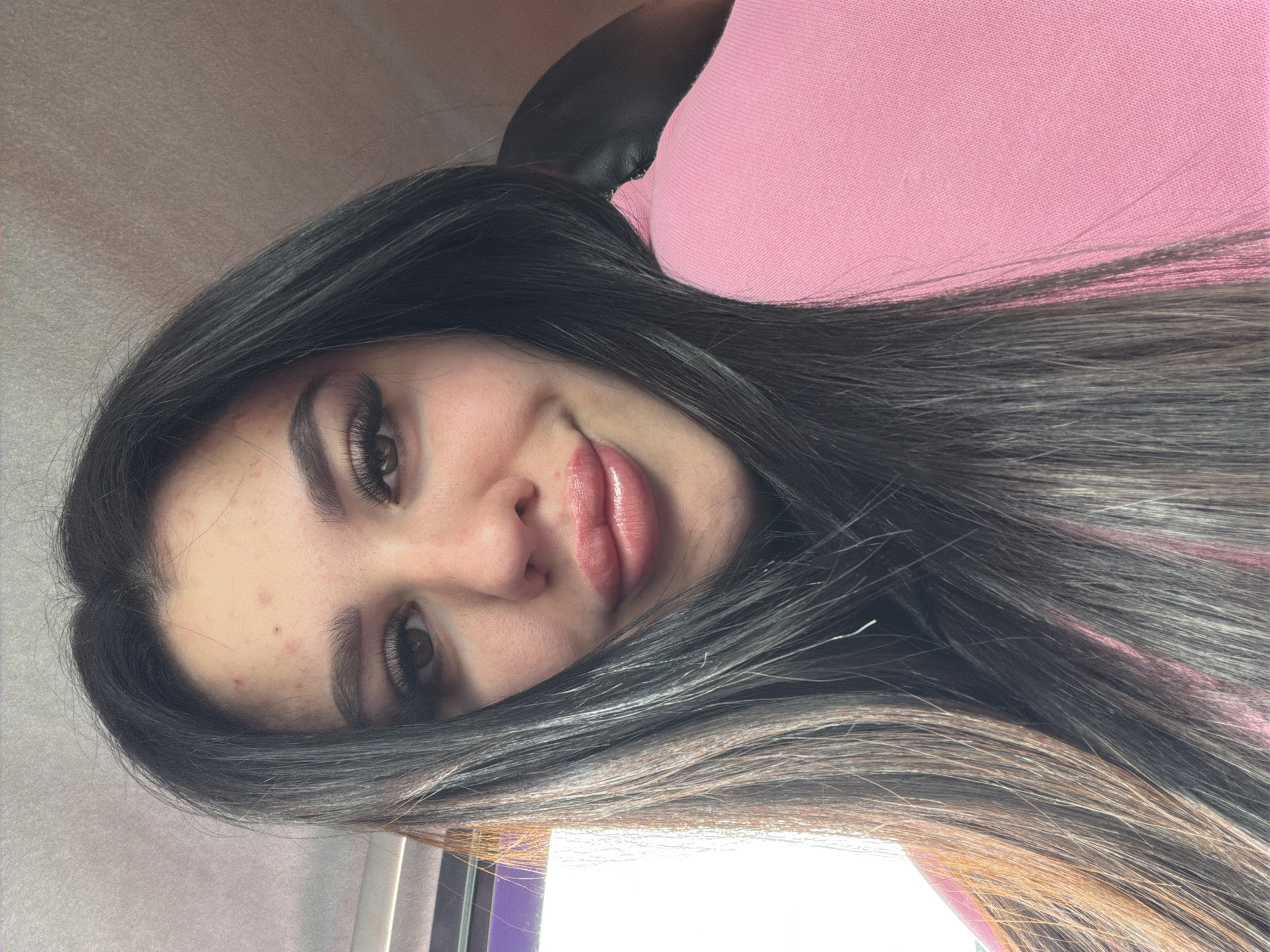 A young woman with long, dark hair, and brown eyes poses for a close-up, rotated portrait. She has fair skin with some visible blemishes on her face. Her lips are accentuated with a glossy lip product. She is wearing a pink top and appears to be seated in a chair with a black headrest. The background is indistinct, but suggests an indoor location, possibly near a window with a purple frame. There is no specific information indicating a location in the city of Фоки, Russia.