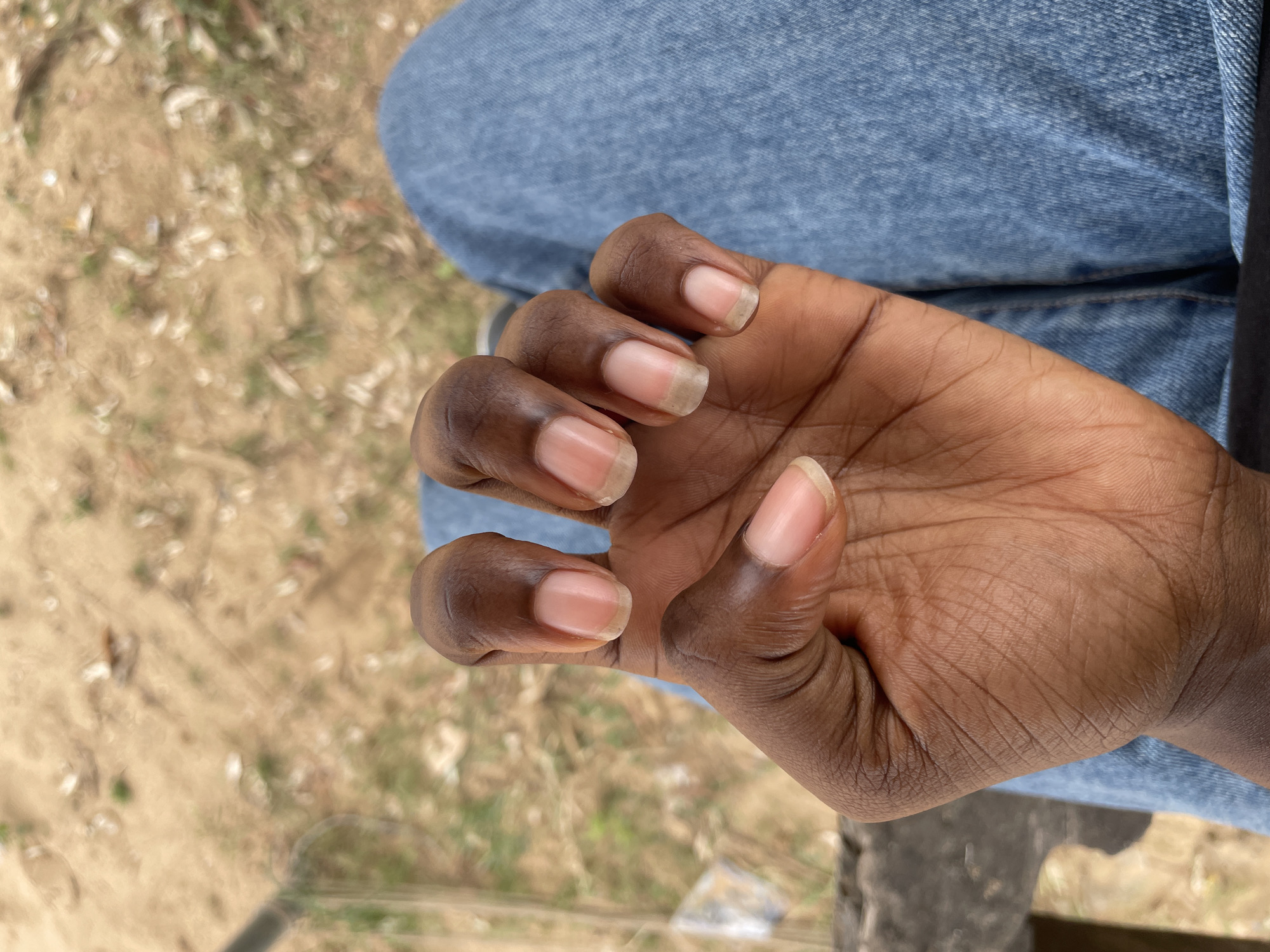 The image shows a close-up view of a person's left hand, with their fingers curled slightly inwards. The hand has dark skin, and the nails appear to be natural, with a light pinkish hue. Some of the nails have a slightly brownish tint at the tips. The person is wearing blue denim jeans, which are visible in the lower right portion of the image. The background is out of focus but appears to be an outdoor setting with sandy ground and some dried leaves or grass. There is a suggestion of a wooden structure on the left side of the frame. The location is identified as Iyafin, Nigeria.