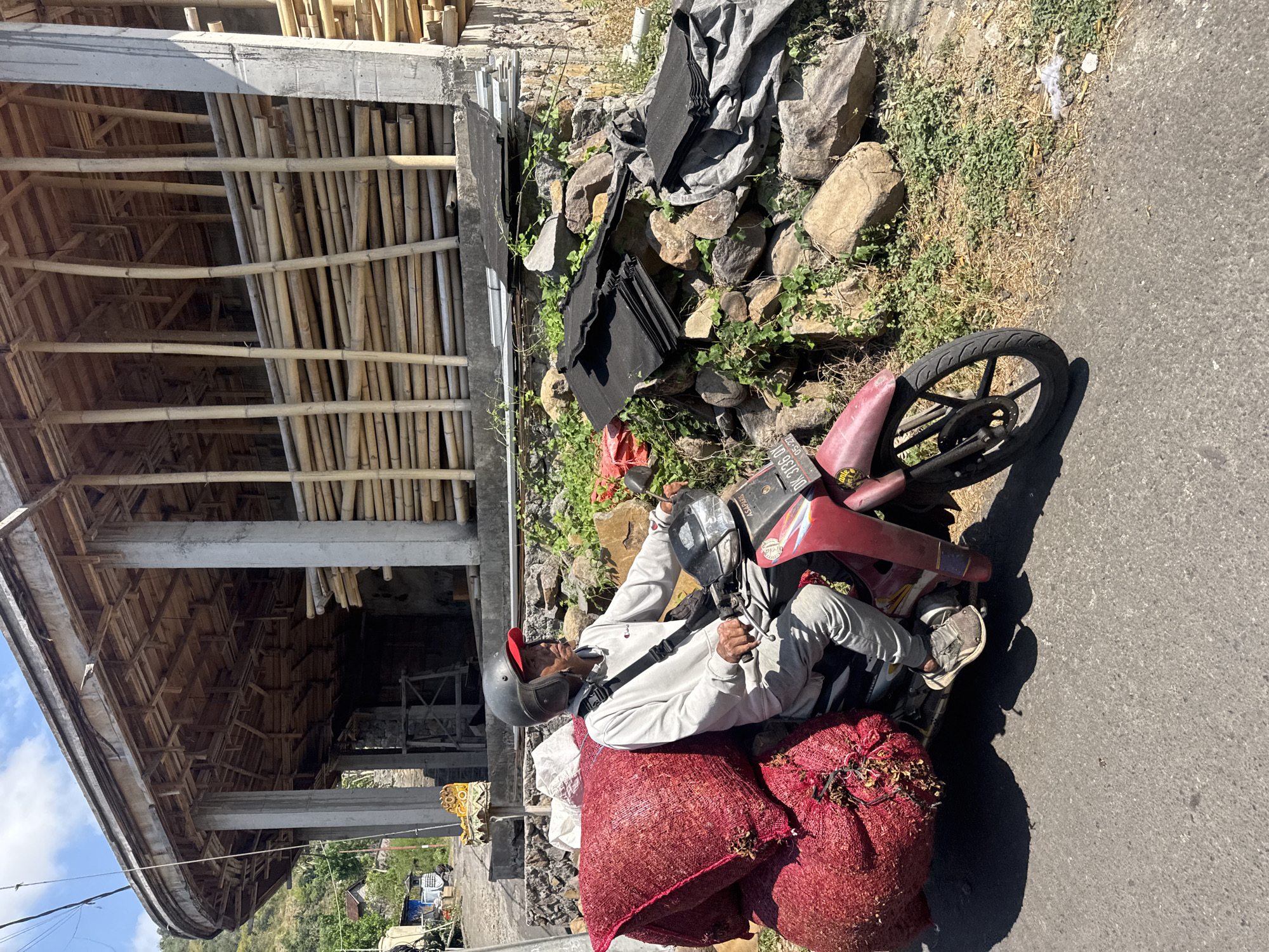 A man wearing a helmet and a light-colored long-sleeved shirt and grey pants rides a red motorcycle on an asphalt road in Abang, Indonesia. The motorcycle is heavily laden with two large, red sacks filled with produce. The man is looking to his right. In the background, a partially constructed building with concrete pillars and a bamboo roof structure is visible. Piles of bamboo poles and dark, flat roofing materials are stacked near the building. Rocks and green vegetation are scattered on the ground along the roadside. The sky is blue with scattered clouds.