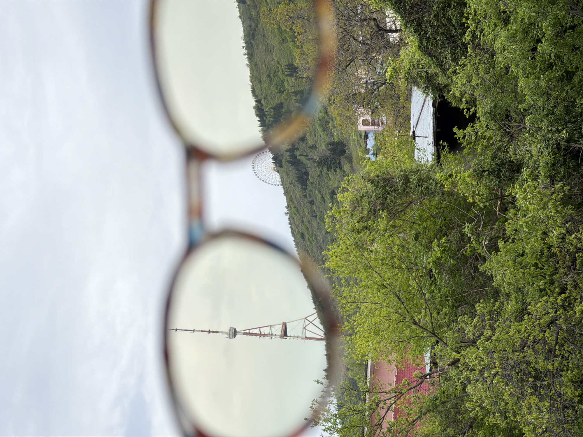 The image is taken through eyeglasses. The upper lens reflects a scene of the Mtatsminda Amusement Park, featuring a Ferris wheel and lush greenery. The lower lens reflects a view of the Tbilisi TV Broadcasting Tower, red structure, and leafy trees. The location is Tbilisi, Georgia.