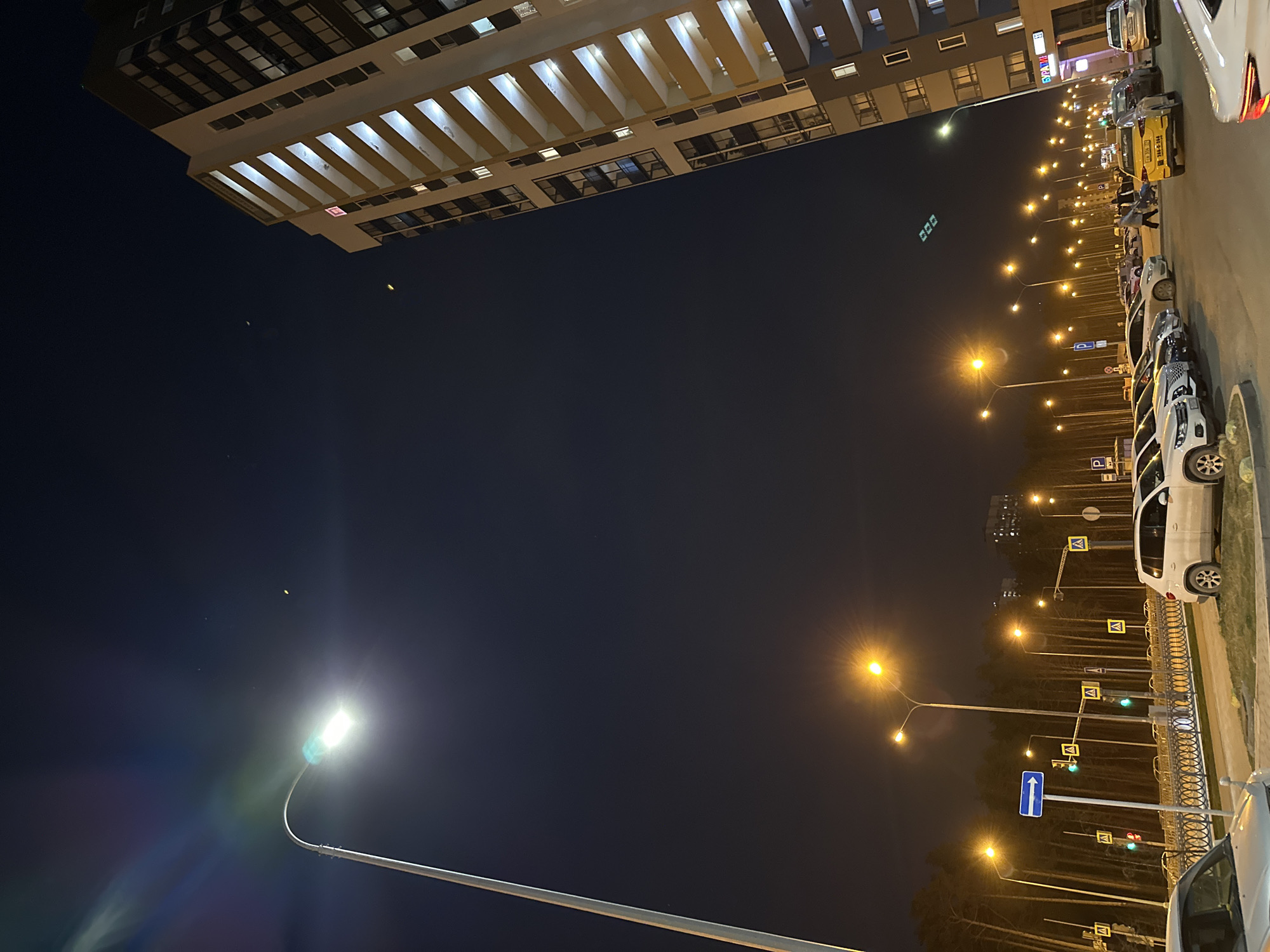 The image shows a night scene in what appears to be a residential or commercial area of Yekaterinburg, Russia. A tall, modern building with illuminated windows dominates the upper left portion of the frame. A row of streetlights lines the right side, illuminating a road with parked vehicles, including cars and what seems to be a taxi. Signage is present, and some greenery or trees are visible along the road. The lighting and architecture suggest an urban environment after dark. No people are clearly visible in the scene.