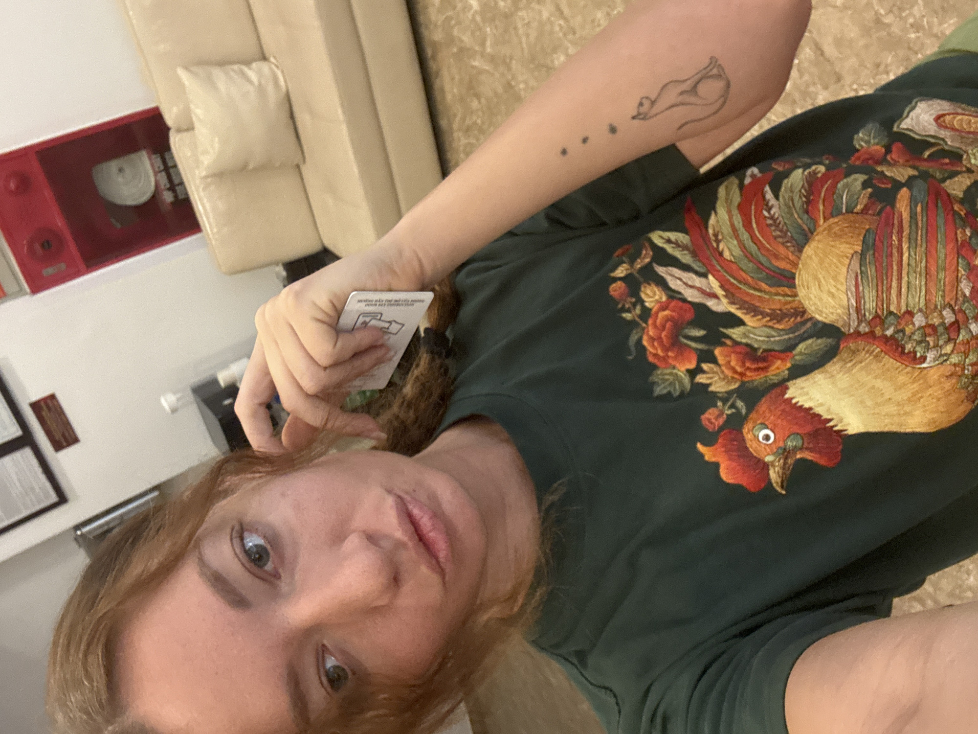 The image depicts a woman in a dark green t-shirt with an embroidered rooster and floral design. She is holding a white card near her face and has a small tattoo of a cat and three dots on her left forearm. In the background, a beige sofa is visible, along with a red fire extinguisher cabinet mounted on a white wall. The scene appears to be indoors, possibly in a hotel lobby or common area in Hanoi, Vietnam, indicated by the presence of the Vietnamese text on the red cabinet.