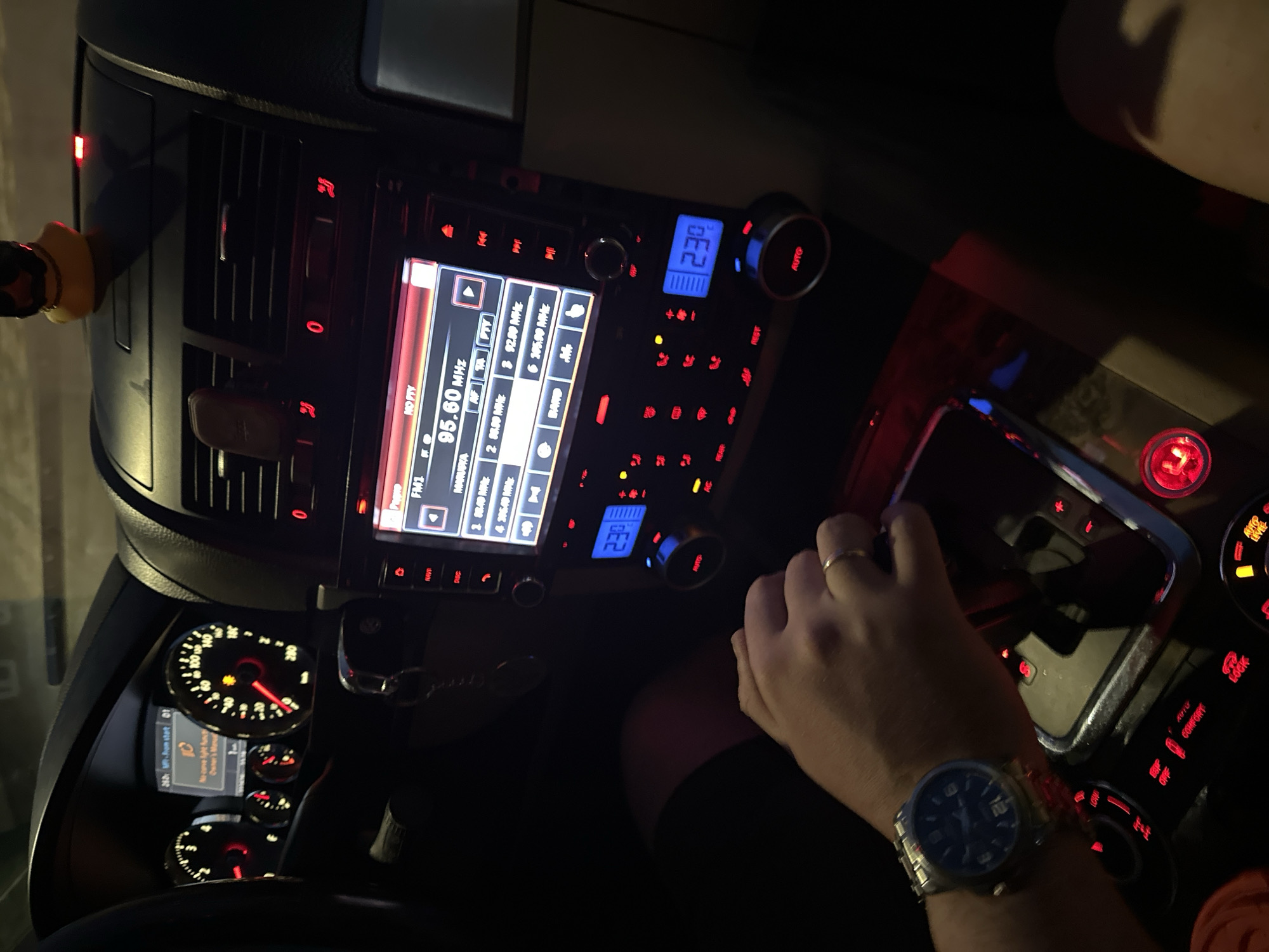 The image shows the interior of a car, with the focus on the dashboard and center console. The car radio display is illuminated and tuned to 95.60 MHz, with other radio frequencies also visible on the screen. The car's speedometer and other gauges are partially visible in the background. A hand wearing a watch and a ring is resting on the gear shift. The overall environment is dark, with the red and blue lights from the car's controls providing some illumination. The text on the screen suggests Russian language radio stations, potentially placing the car in Russia. There is no direct evidence of the location being in Nizhny Novgorod.