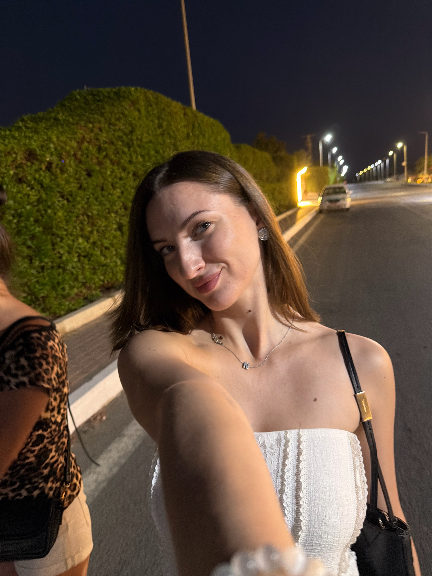 A young woman with dark hair and fair skin takes a selfie at night on a paved street in Sharm El Sheikh, Egypt. She is wearing a white strapless top with lace trim and a delicate silver necklace. Her left arm is extended, holding the camera. To her left, another person wearing a leopard-print top and white shorts is partially visible. The street is lined with streetlights, and a car is parked on the right side of the road. Lush green bushes border the left side of the street. The sky is dark blue, indicating it is nighttime.