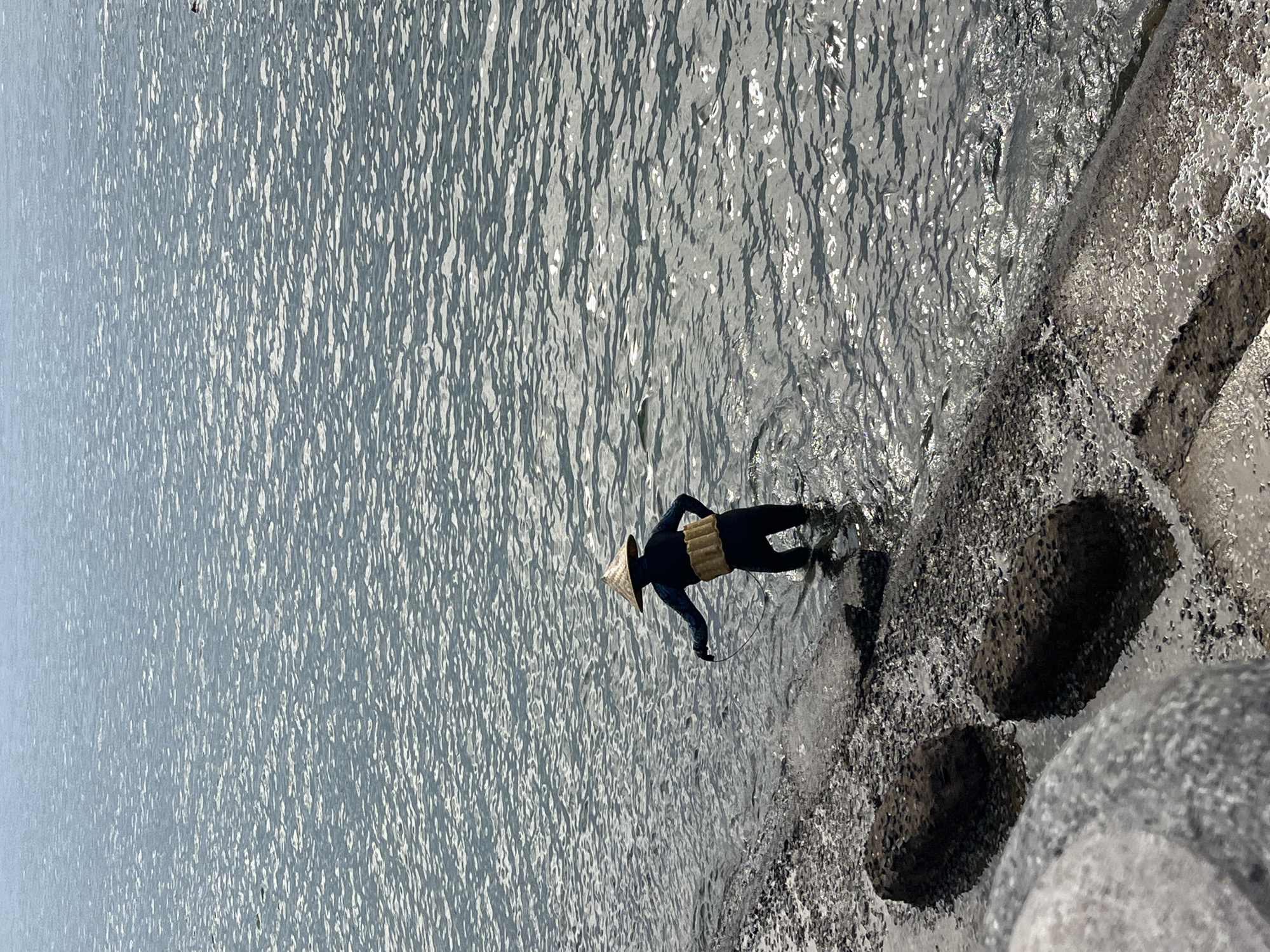A person in a black suit, a conical hat, and a yellow floatation device stands waist-deep in shimmering water, next to rocky formations with holes, likely located in or near Bac Nha Trang Ward, Vietnam. The scene appears to be photographed outdoors.