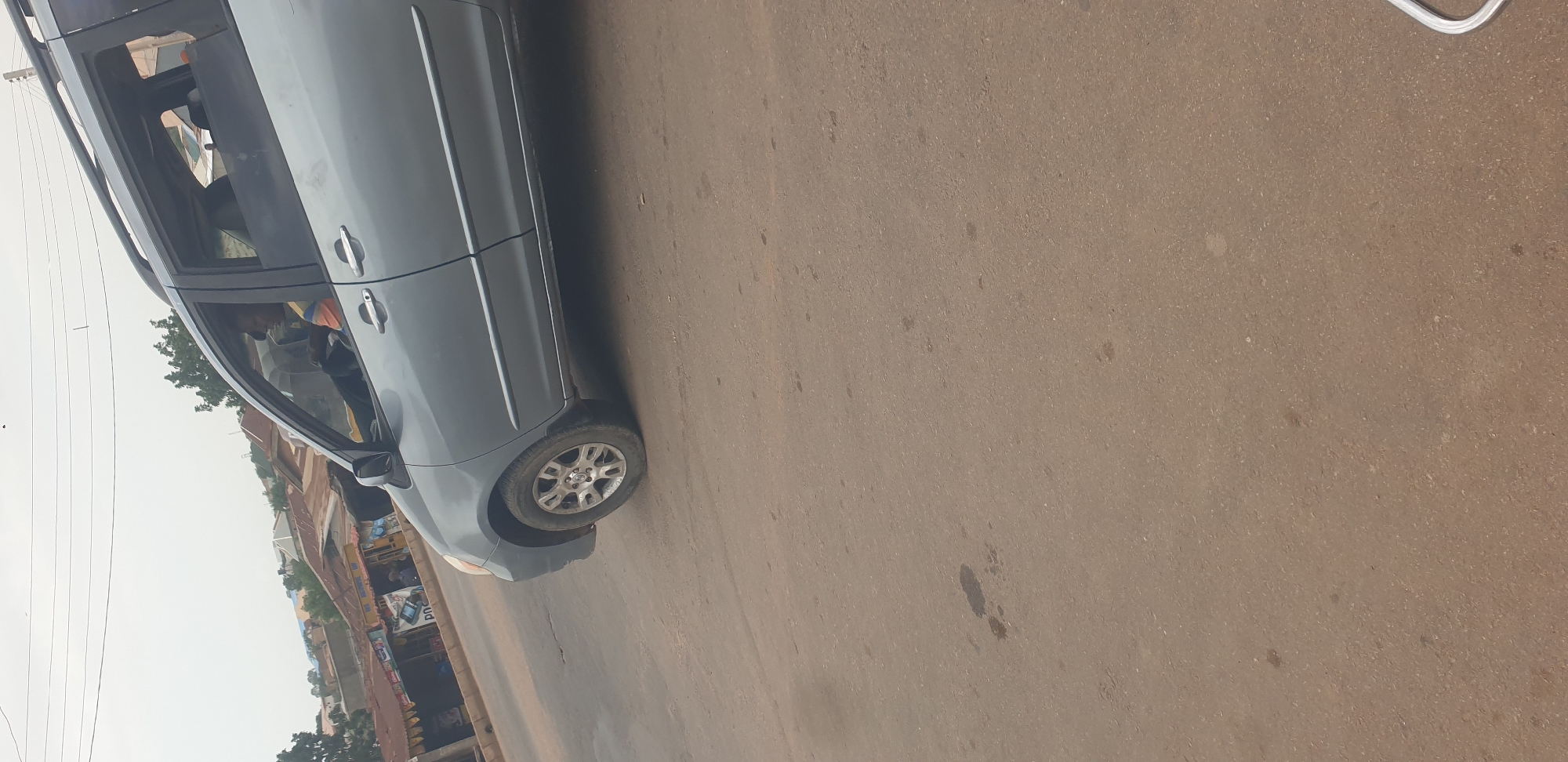 A grey minivan is parked on the side of a paved road in Barakin Lerawi, Nigeria. The minivan is facing the viewer's left. The driver, a man wearing a blue and orange shirt, is visible through the driver's side window, looking down at something in his hands. The scene appears to be daytime, with overcast skies suggesting a cloudy or possibly rainy day. The road is a light brown asphalt with some dark spots, indicative of dirt or oil stains. In the background, on the left side of the image, there are some low-rise buildings, and trees can be seen. The architecture of the buildings suggests a commercial area, with what look like shops lining the street. Power lines are also visible running overhead. There is no discernible text in the image.