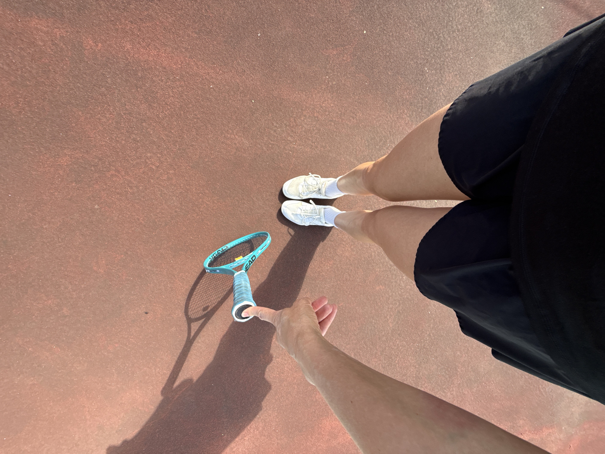 A first-person perspective from a person standing on a tennis court, with their left arm extended and pointing towards a tennis racket. The racket, with a teal frame and grip, lies on the reddish-brown court. The person is wearing white athletic shoes, white socks, and black shorts. A shadow of the racket is cast on the court to the left. The scene appears to be outdoors in daylight. The location in the city of Chaikovsky, Russia, is not evident from the image.
