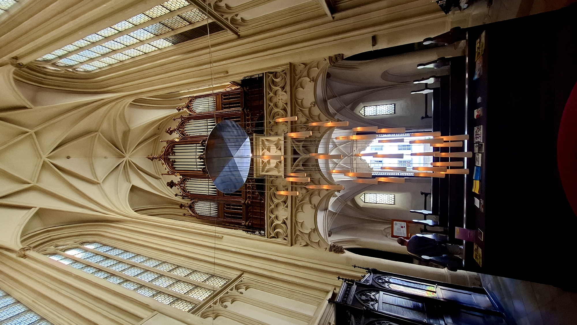 An interior shot of the Augustinian Church (Augustinerkirche) in Wien, Austria, captures a dramatic upward view of the nave. The focal point is a large, modern chandelier with numerous cylindrical lights descending from a circular disk suspended from the ceiling. Behind the chandelier, an ornate pipe organ is situated in an upper gallery. The church's architecture features high, vaulted ceilings with intricate ribbed patterns and tall, arched windows with leaded glass panes, characteristic of Gothic style. The walls and columns are rendered in a light, neutral stone. In the foreground, a dark-colored table displays a variety of brochures and books. To the left, a man wearing a dark jacket and gray pants stands facing away from the camera, looking towards the area with the table and benches. Benches are visible on either side of the central aisle, with a few small windows providing light from the upper sections of the church. The overall lighting is subdued, with the chandelier and windows providing the primary illumination.