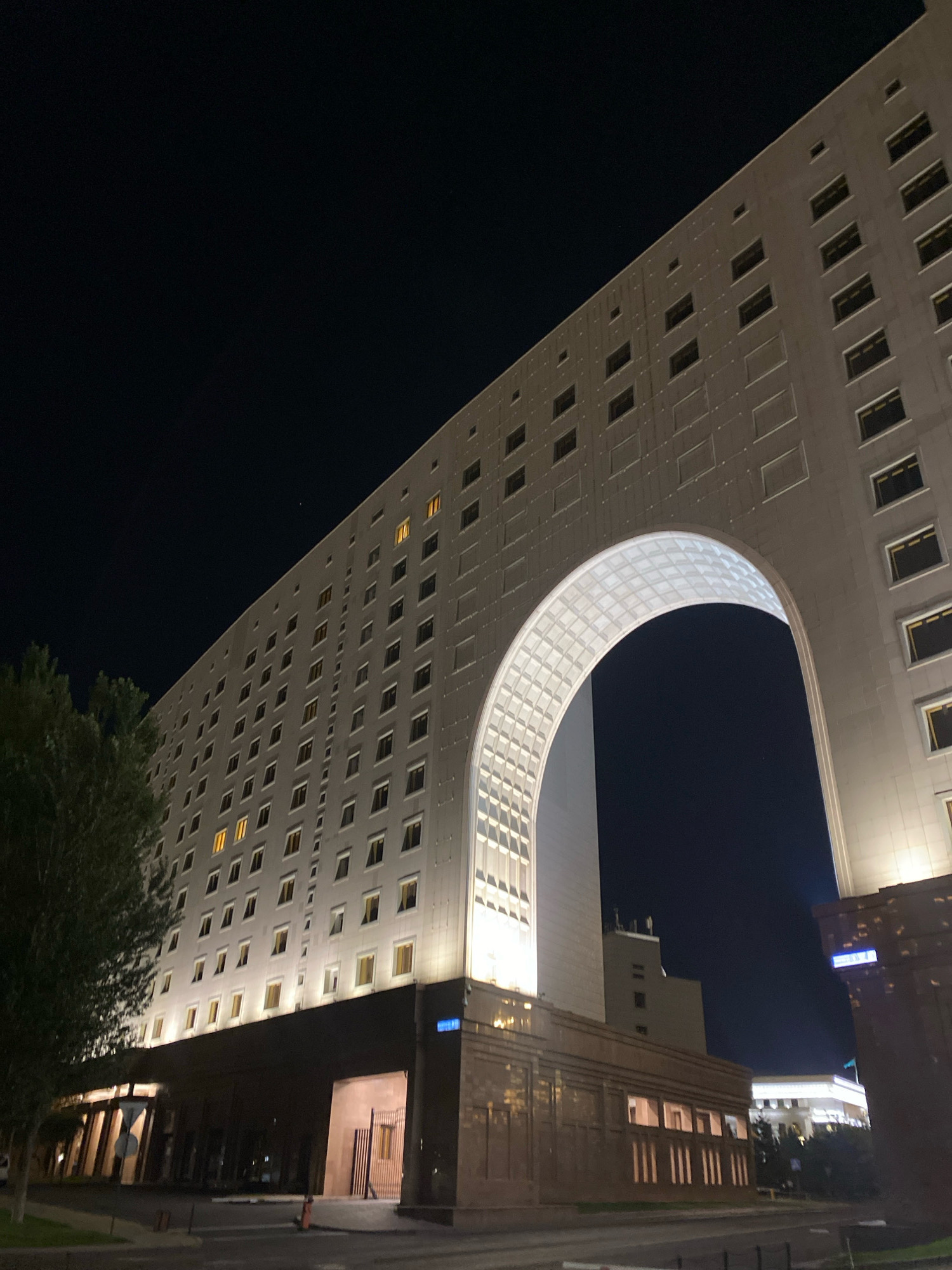 The image shows a night view of a large, modern building in Astana, Kazakhstan. The building is tall and imposing, with a striking architectural design featuring a massive arched opening. The facade is light-colored and dotted with numerous windows, many of which are illuminated with a warm yellow light, suggesting occupancy. The archway itself is also brightly lit, with a patterned interior that resembles a ribbed vault.
Below the main structure, a darker, lower section of the building is visible, featuring a row of vertically oriented windows that also emit light. To the left, a large, leafy tree stands in front of the building, its dark silhouette contrasting with the illuminated architecture. In the background, other buildings and streetlights are faintly visible against the dark night sky. The overall impression is one of urban grandeur and nighttime activity. The building appears to be a hotel or an office complex.