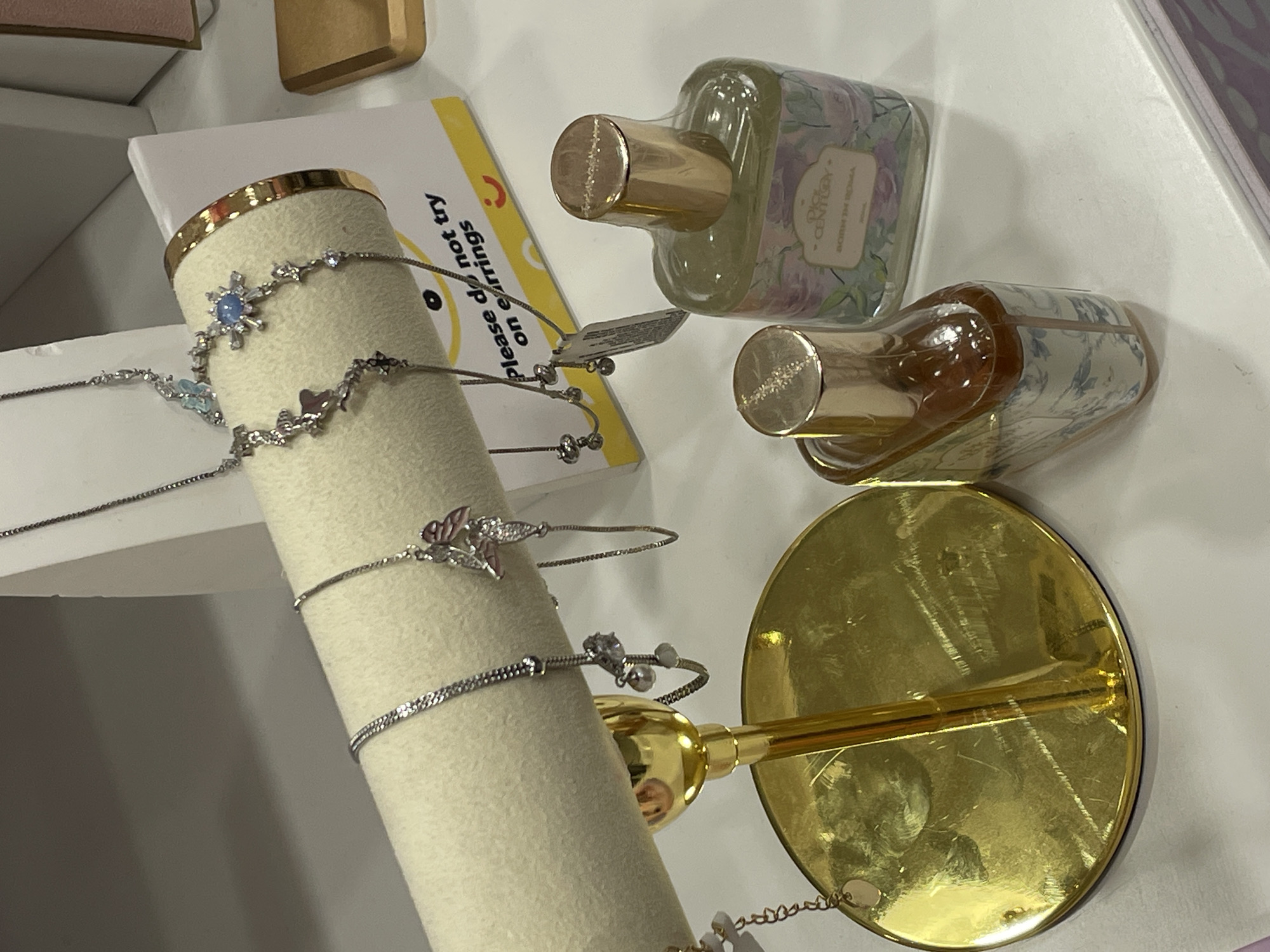 This is a close-up, eye-level shot of a retail display, likely in a department store or boutique, potentially located in Davao City, Philippines. The scene is brightly lit and shows a white, cylindrical display stand for jewelry, covered in a light-colored fabric. Hanging from the stand are several delicate silver necklaces, adorned with charms that appear to be small butterflies and celestial elements. One necklace features a blue stone surrounded by small clear stones, resembling a snowflake. To the right of the jewelry display, a yellow sign with black text reads, "Please do not try on earrings," accompanied by a smiley face graphic and a hint of a red border.
In the foreground, two glass perfume bottles with gold caps are visible on a white surface. The bottle on the right contains a pale green liquid and has a label with floral imagery and text that appears to read "DICE CENTURY" and "BORN IN ROMA". The bottle on the left contains a darker amber liquid and has a label with a blue and white floral pattern. A polished gold disc-shaped stand supporting the jewelry display is also prominent in the lower left quadrant of the image, showing some scuff marks. The background is out of focus, showing a muted grey wall. There are no people visible in the image.