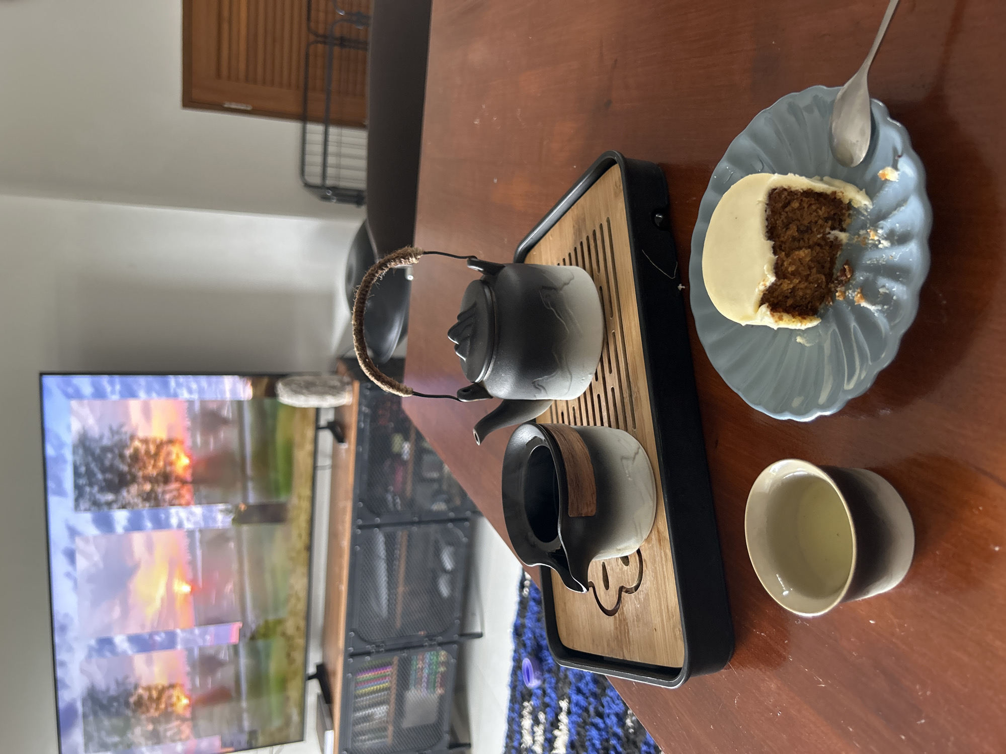 The image depicts a still life on a wooden table, likely in a residential setting in Chang Phueak, Thailand, given the context provided. In the foreground, a slice of carrot cake with white frosting sits on a light blue scalloped plate, accompanied by a silver spoon. To the left, a small ceramic teacup contains a pale green liquid. Central to the image is a traditional Chinese tea set on a bamboo tray. This includes a black and white gradient teapot with a wicker handle and a matching gaiwan, used for brewing tea. Behind the tea set, a large flat-screen television displays a vibrant sunset scene with silhouetted trees. The background reveals a modern entertainment console with metal mesh doors, and further back, a louvered wooden door and a metal rack. The overall ambiance suggests a moment of relaxation and enjoyment, possibly in the evening.